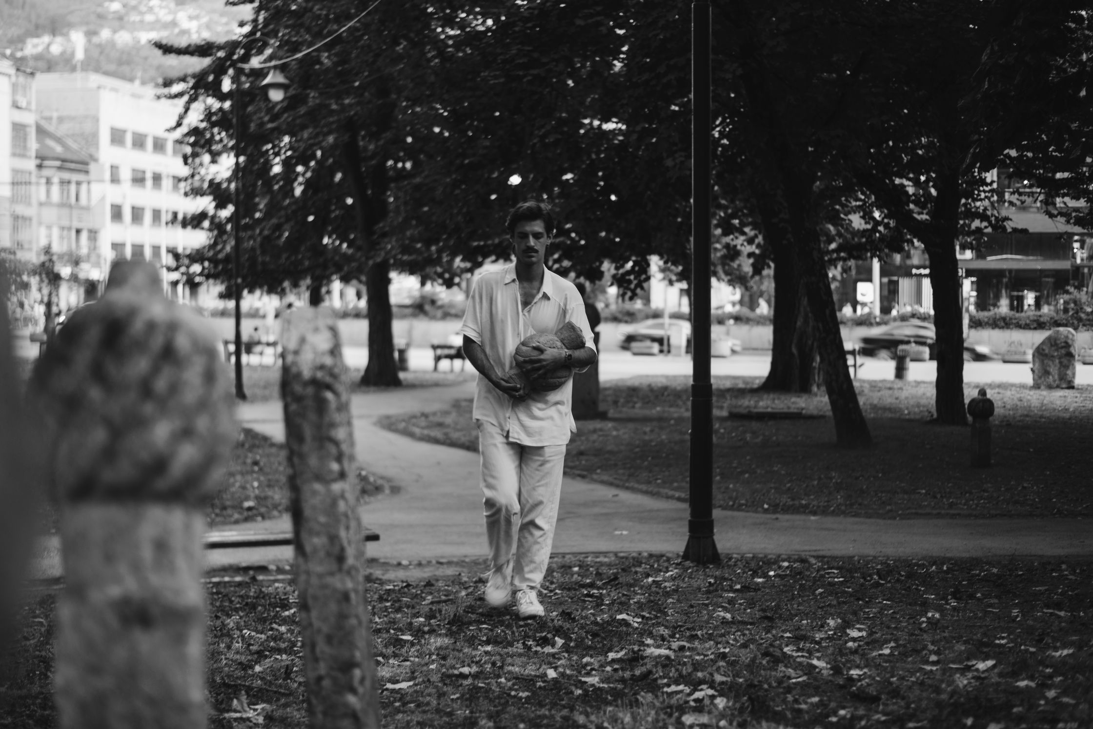 A man in a light-colored outfit walks through a park holding a ball in his arms.