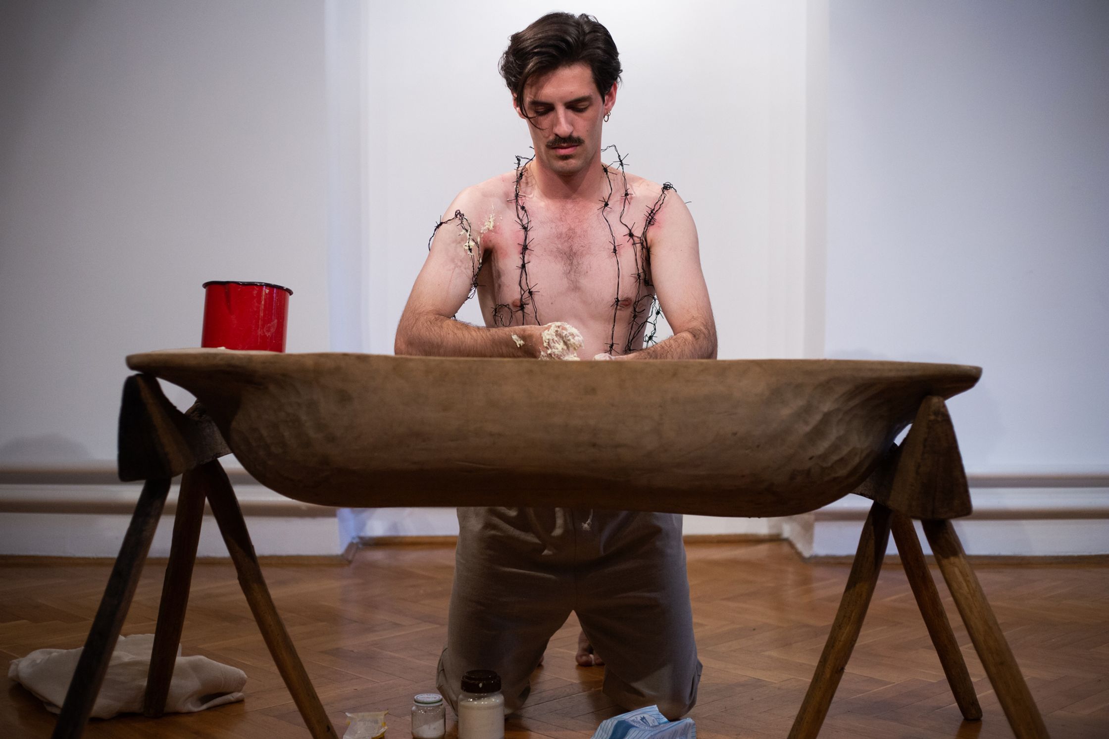 A barefoot man kneels before a wooden trough, kneading dough, while barbed wire is intertwined into his body and a red pot sits on the table.