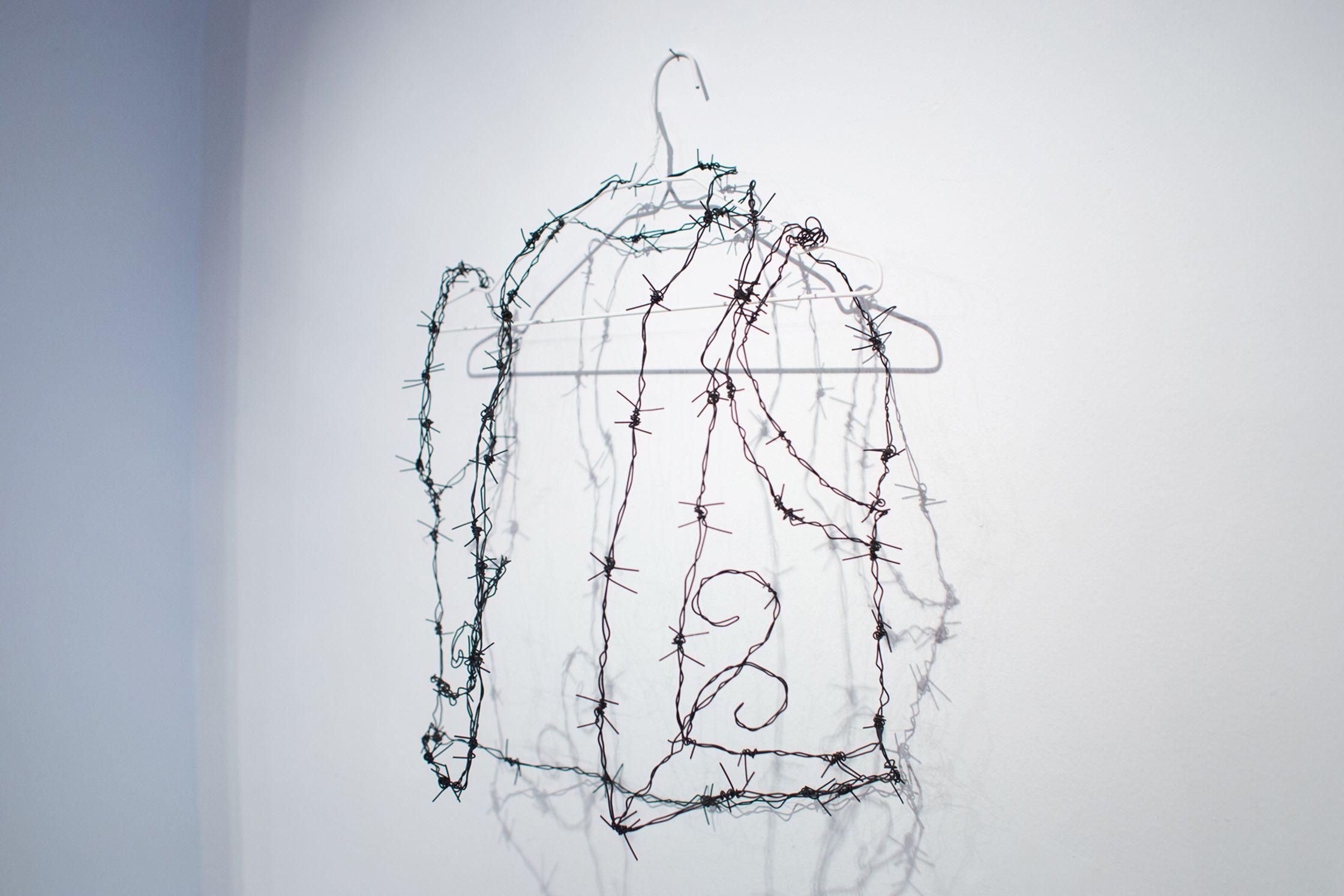 A delicate shirt-shaped sculpture made of barbed wire hangs on a white wall.