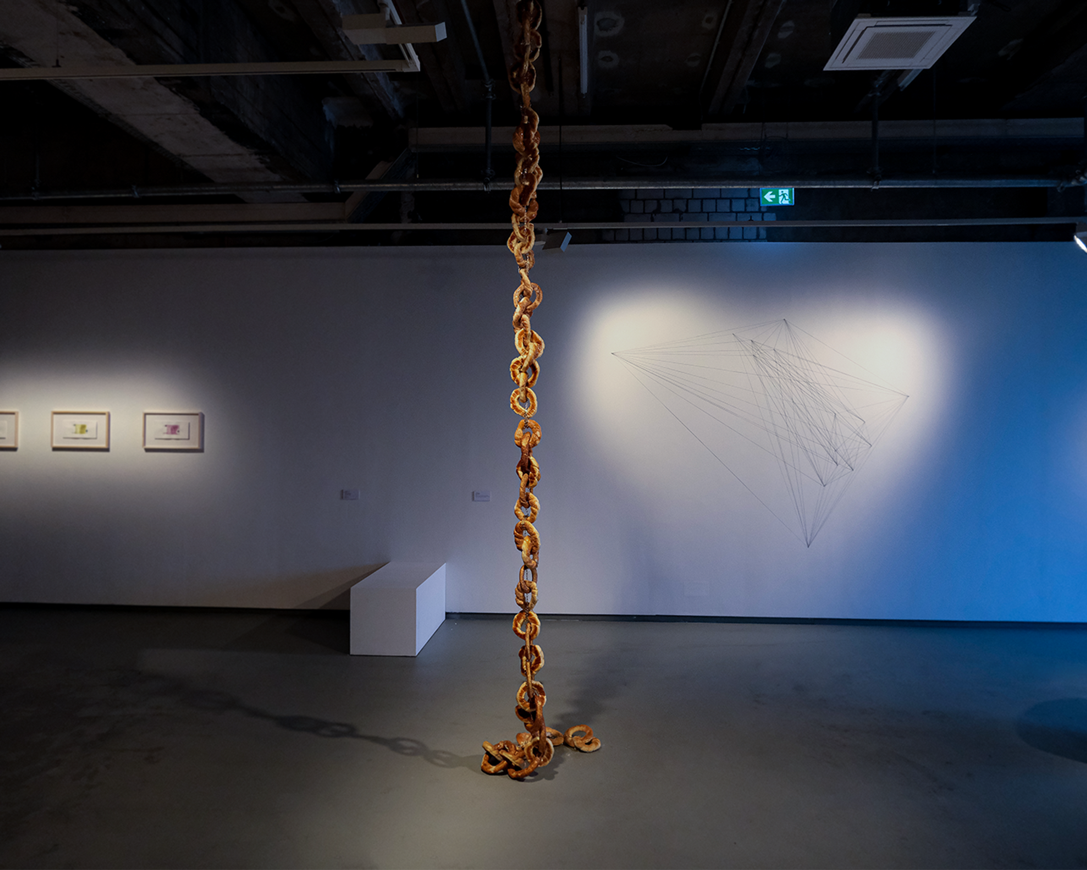 Indoor art installation featuring a large, ornate golden chain extending from the ceiling to the floor, with geometric string art on the wall in the background.