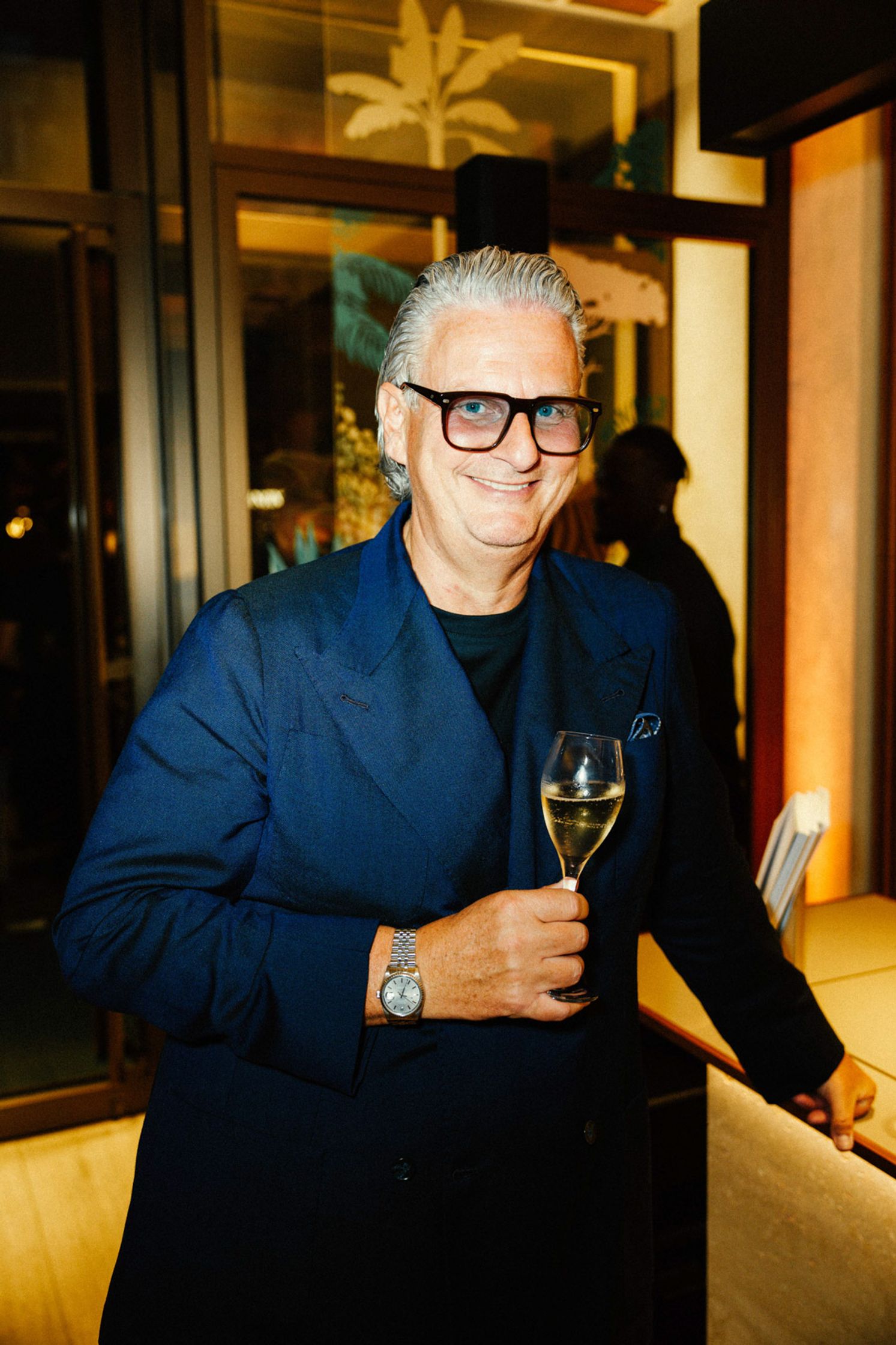 Eleganter Mann mit grauem Haar, Brille und dunklem Sakko hält ein Champagnerglas, Rolex-Uhr am Handgelenk, lächelnd beim Rolex Boutique Berlin Event