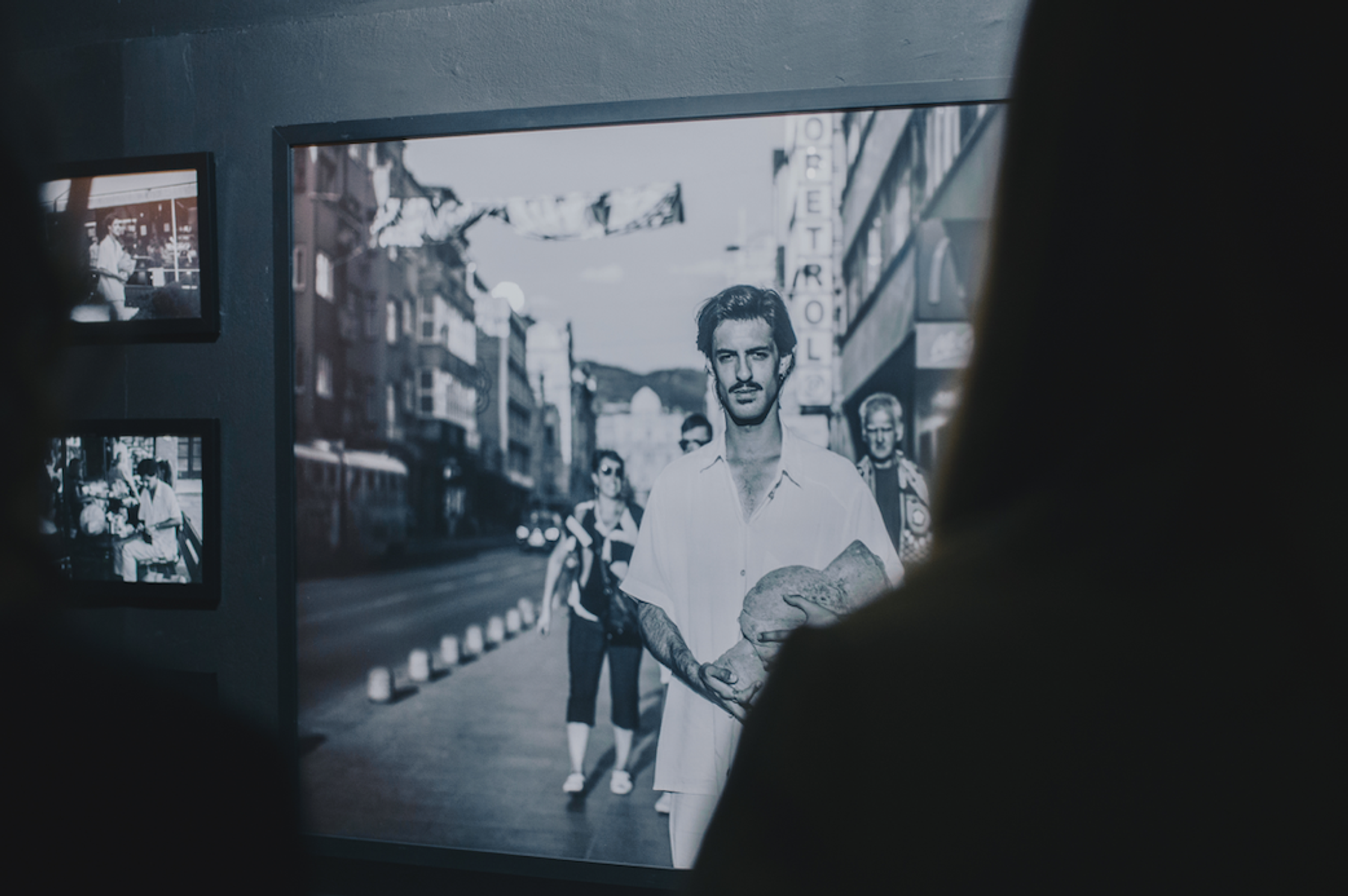 A black and white photo of a man holding a large rock on a street is displayed on a wall with two smaller photos to the left, partially obscured by a person's silhouette.