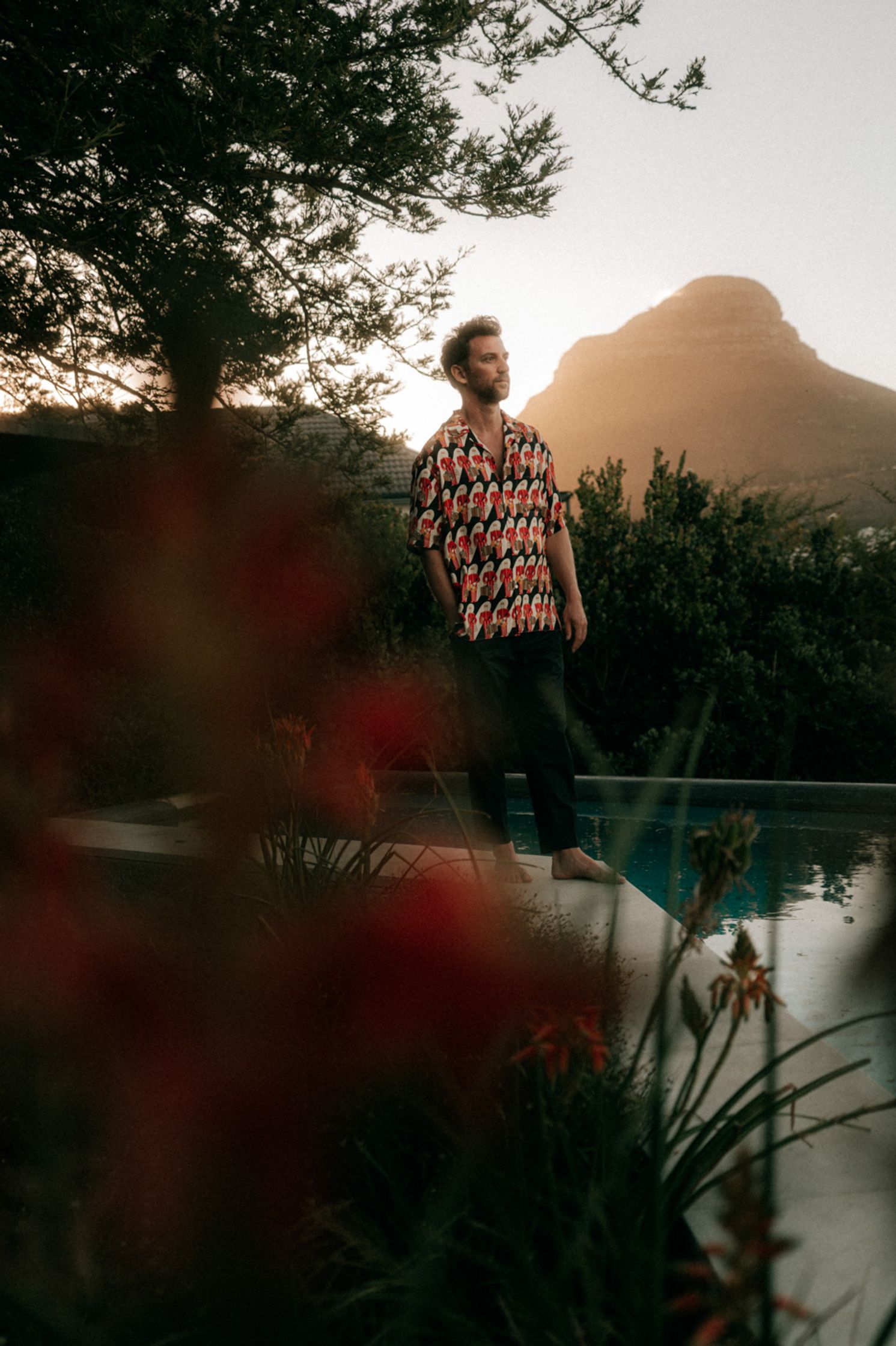 Künstler Paul Schrader steht barfuß am Rand eines Infinity-Pools in einer Kapstädter Villa, gekleidet im bunten Figurenhemd, mit dem Lion's Head im goldenen Abenddunst im Hintergrund