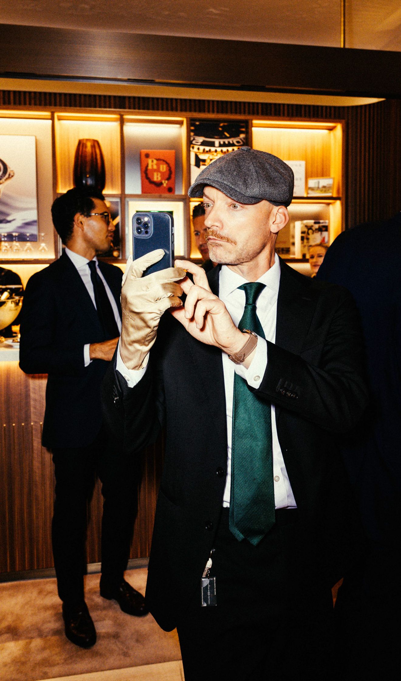 Zwei Männer im Anzug bei einem Event in der Rolex Boutique Berlin: Mann mit Schiebermütze und grüner Krawatte schaut aufs Smartphone, Uhren-Vitrinen und gerahmte Fotos im Hintergrund
