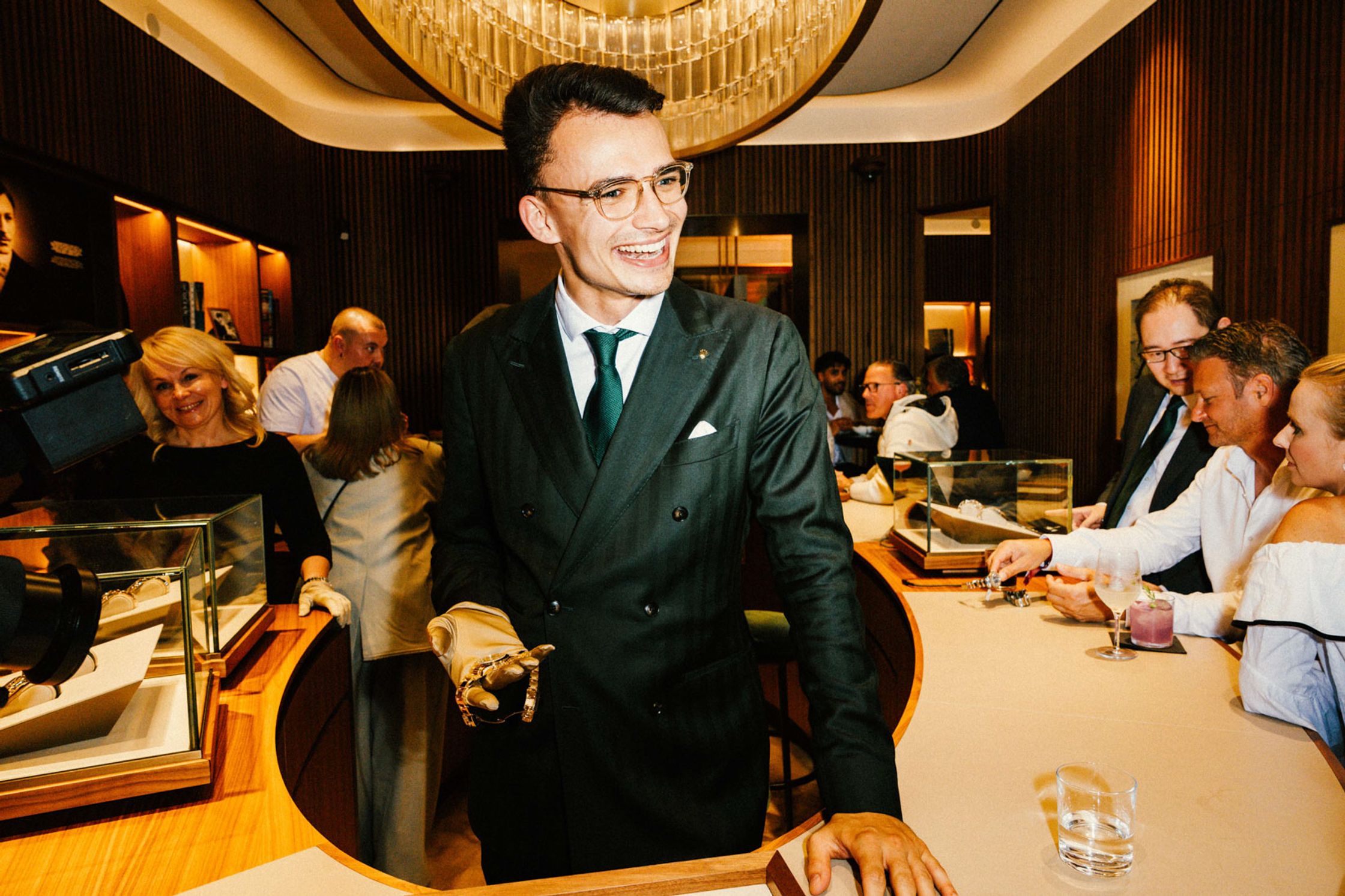 Junger Rolex-Mitarbeiter im dunklen Zweireiher-Anzug mit grüner Krawatte präsentiert lächelnd eine Uhr, gäste im Hintergrund, eleganter Kronleuchter, Rolex Boutique Berlin Event