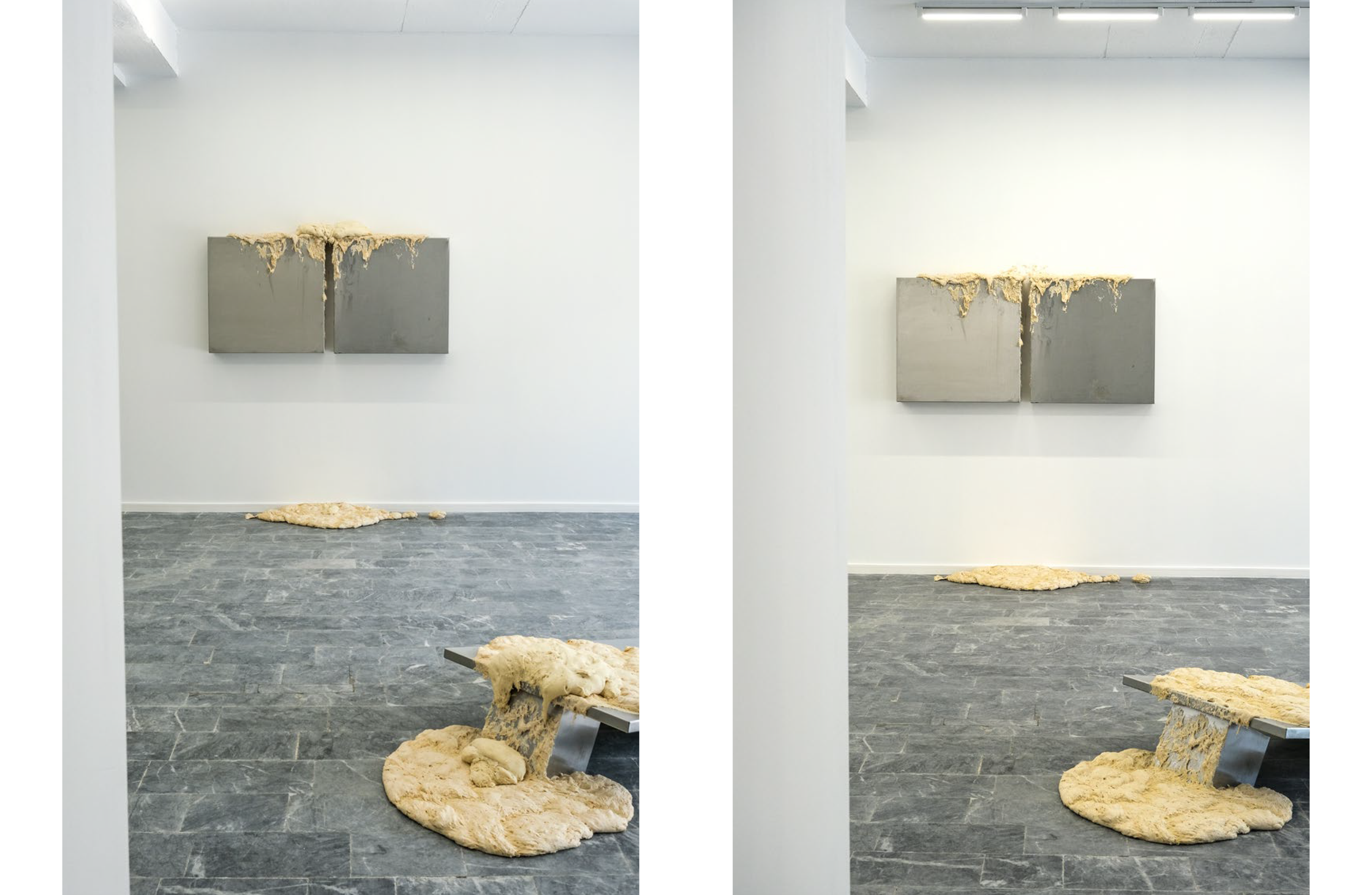 An exhibition space featuring two dark panels on a white wall with light-colored dough-like material spilling from them onto the floor and pooling underneath a bench.