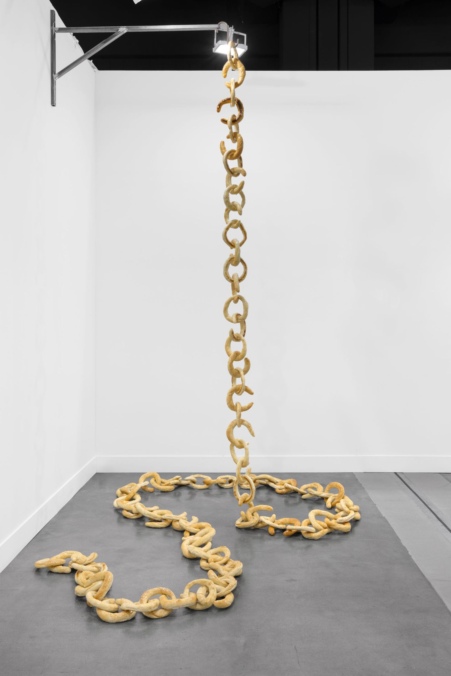 A long chain made of pretzels hangs from a wall-mounted bracket, with the bottom portion resting on the floor in a scattered pattern.