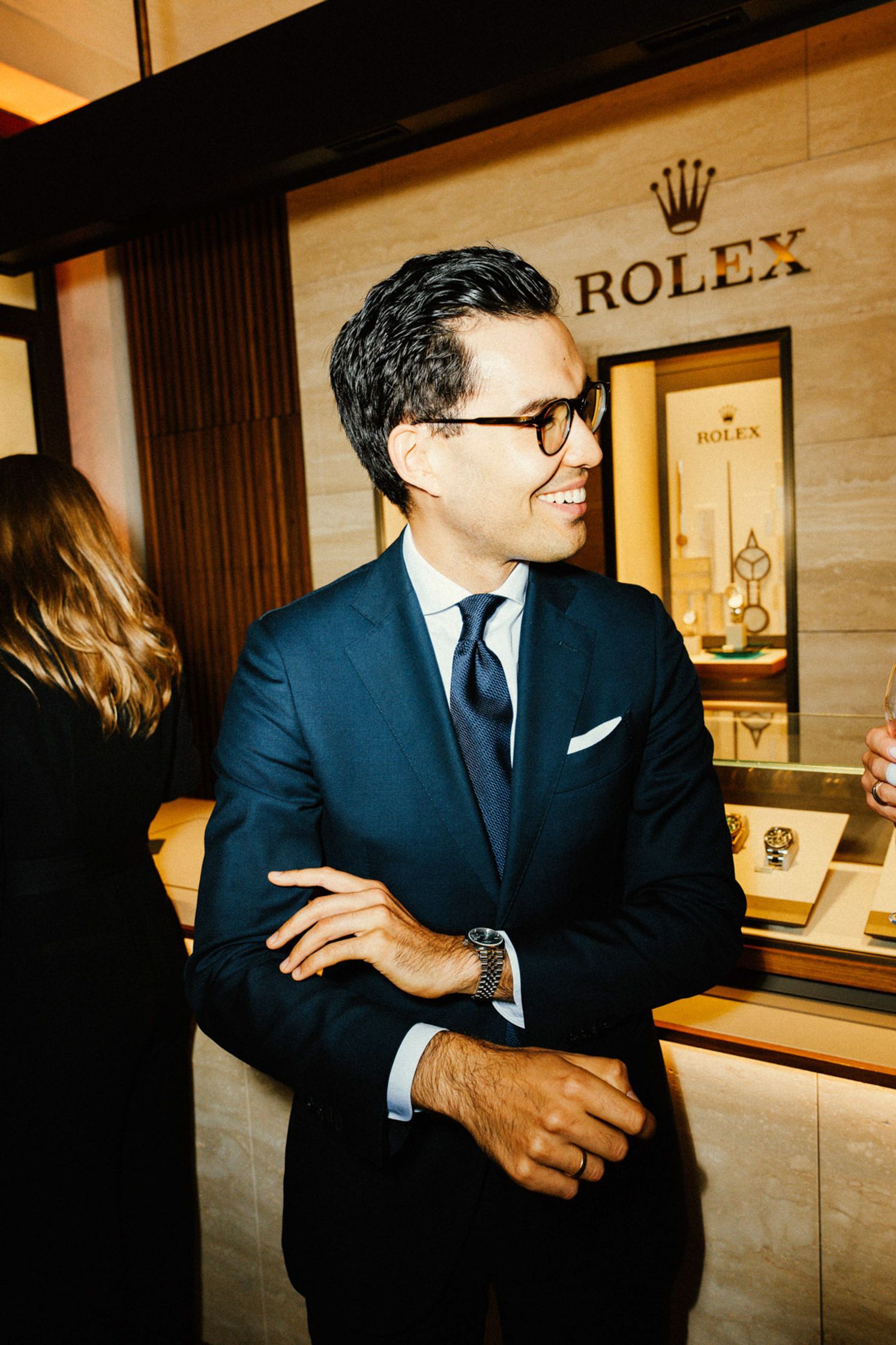 Eleganter junger Mann im dunkelblauen Anzug mit blauer Krawatte und Brille lächelt beim Rolex Boutique Event Berlin vor beleuchteter Uhren-Vitrine mit Rolex-Logo im Hintergrund