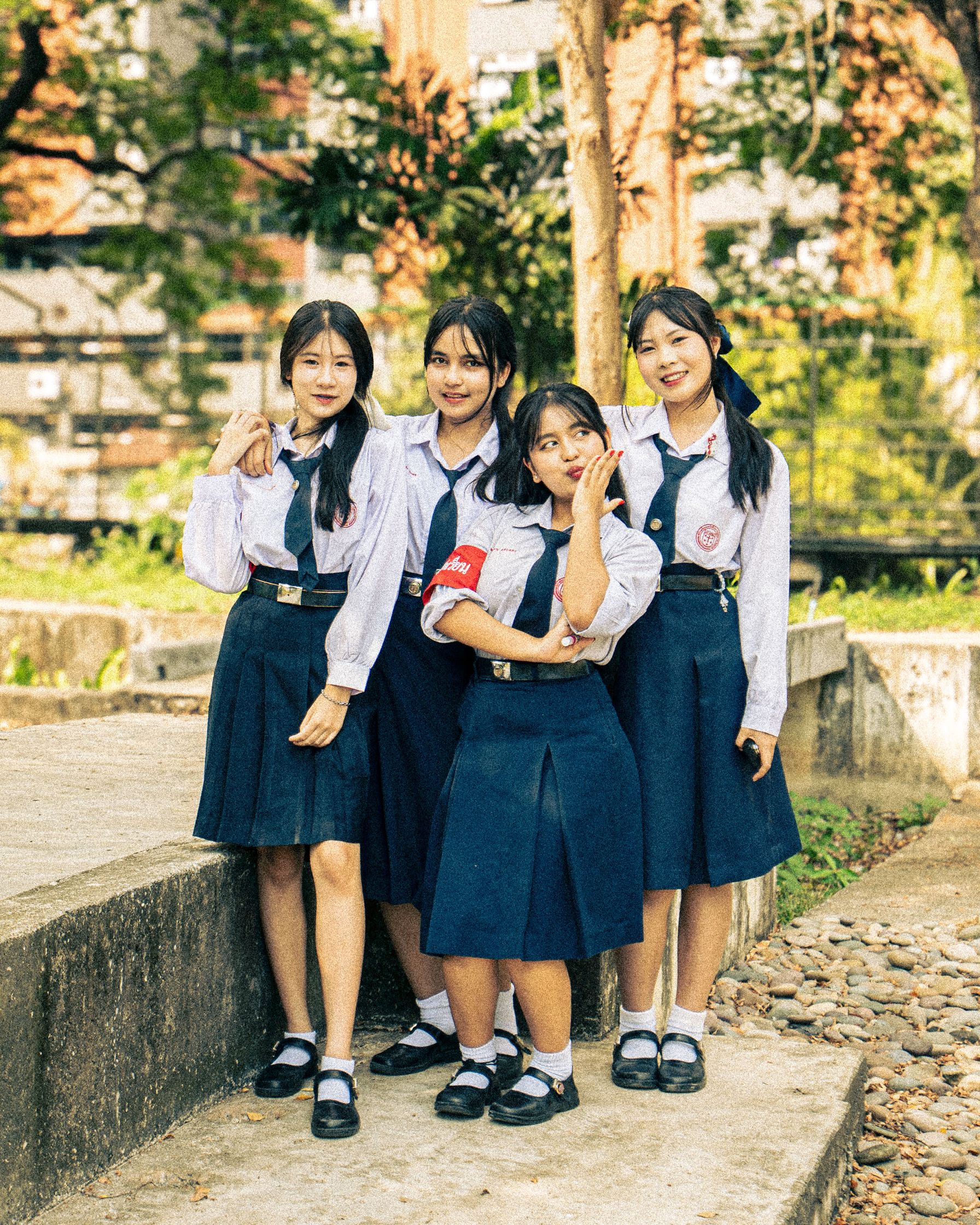 A candid portrait of a group of Thai schoolgirls in uniform sharing laughter in a public park. Capturing the youthful energy within Bangkok's urban sprawl.
