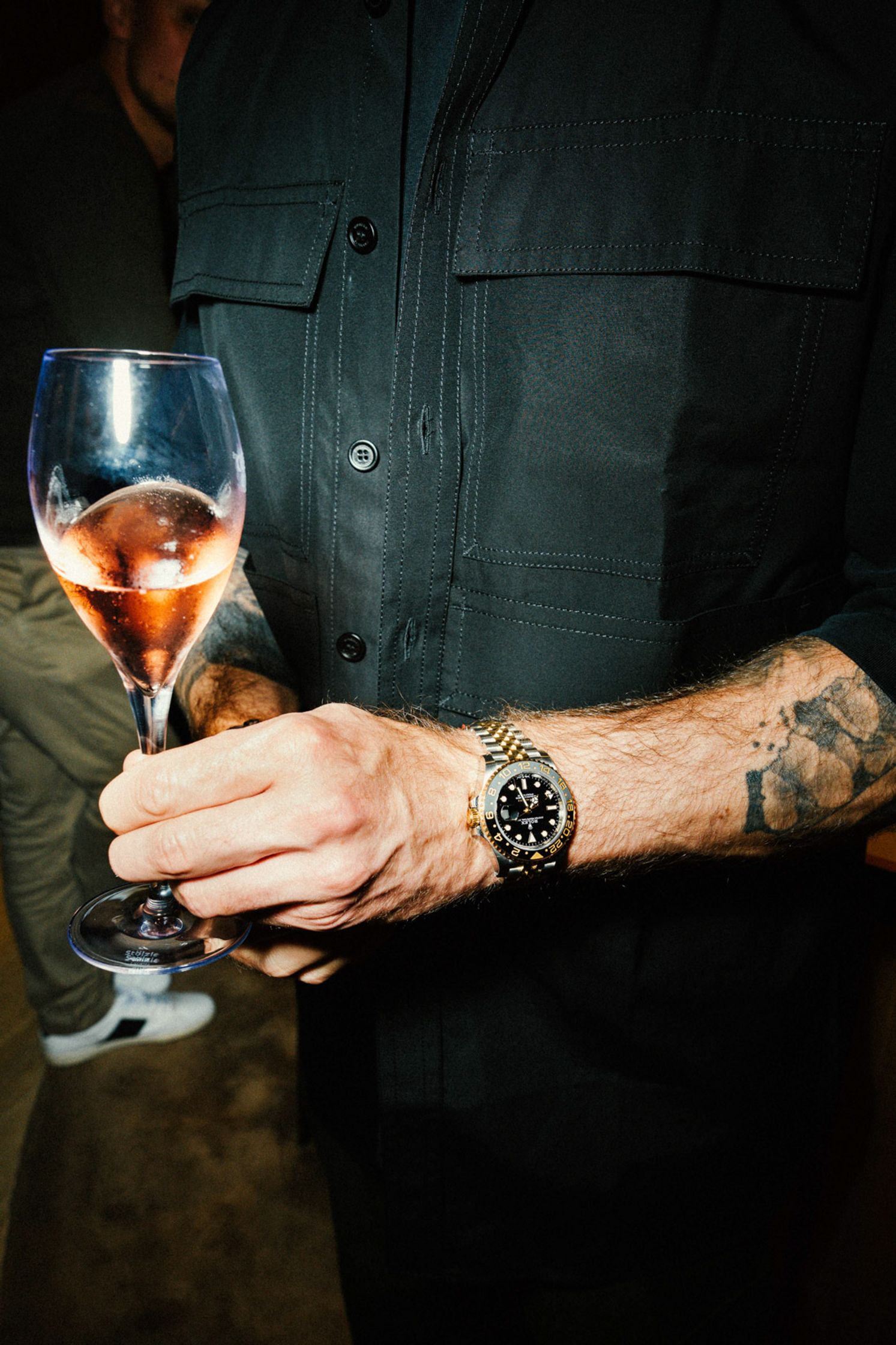 Nahaufnahme eines tätowierten Handgelenks mit Rolex GMT-Master II in Gold/Stahl beim Rolex Boutique Event Berlin – Hand hält ein Rosé-Champagnerglas