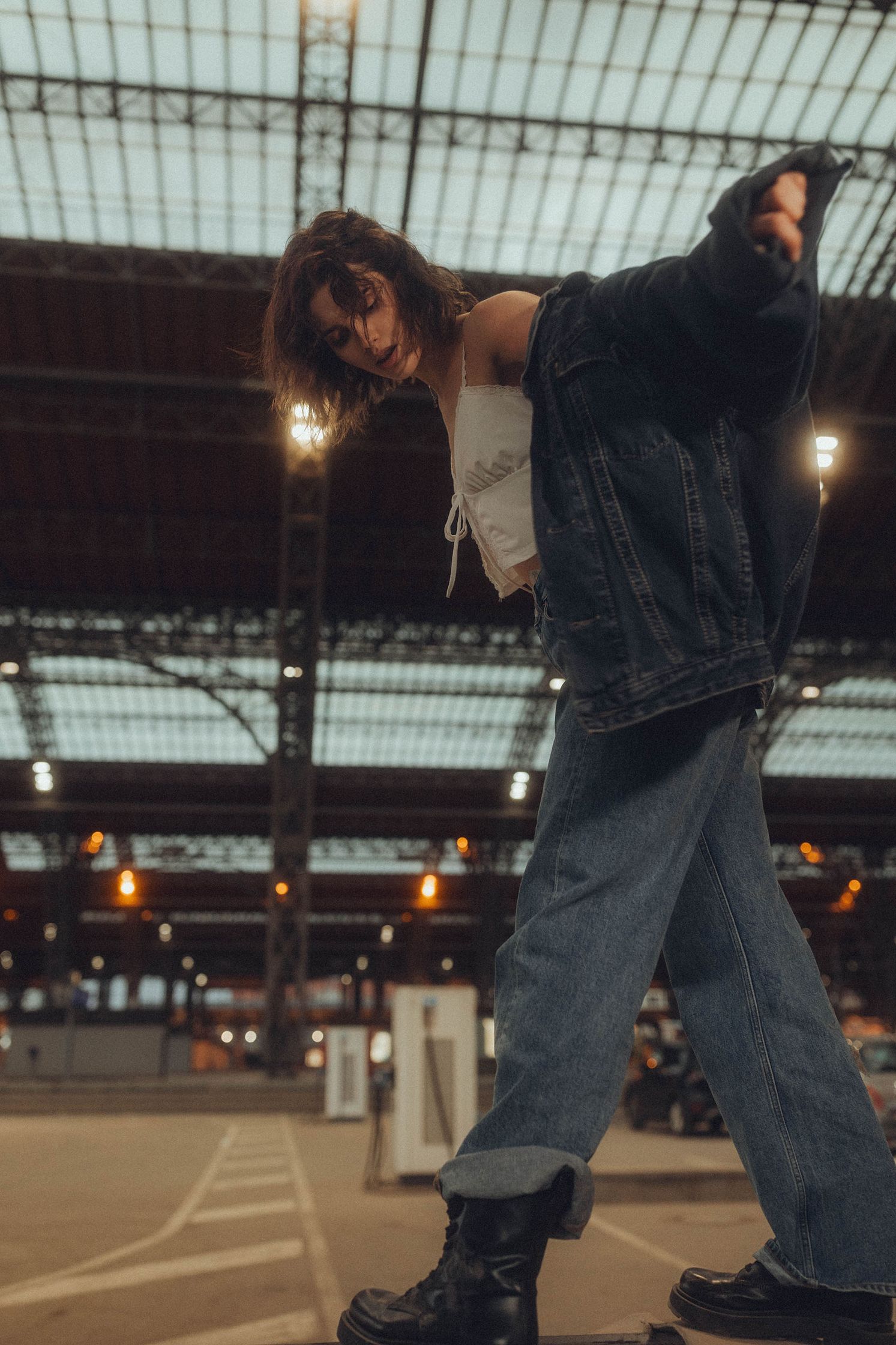 A young woman in a white top, denim jacket, and jeans bends over, looking up at the viewer, under a glass ceiling.
