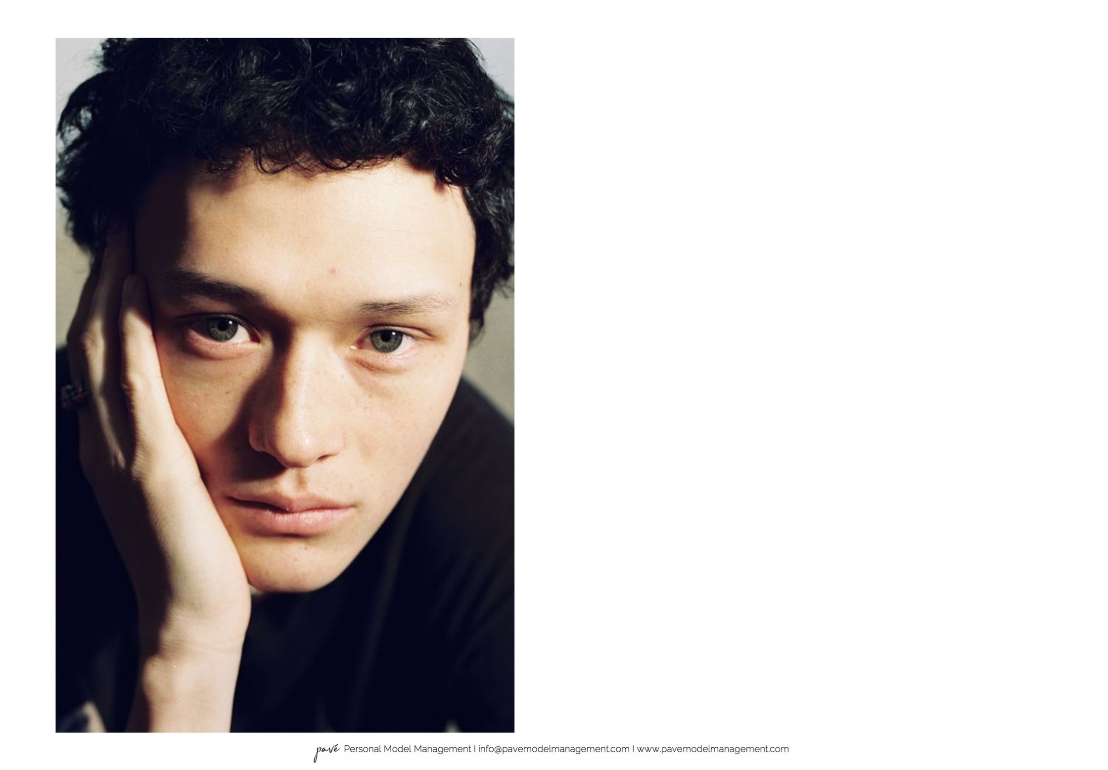 A dark-haired person with light eyes gazes directly at the viewer, resting their chin on their hand.