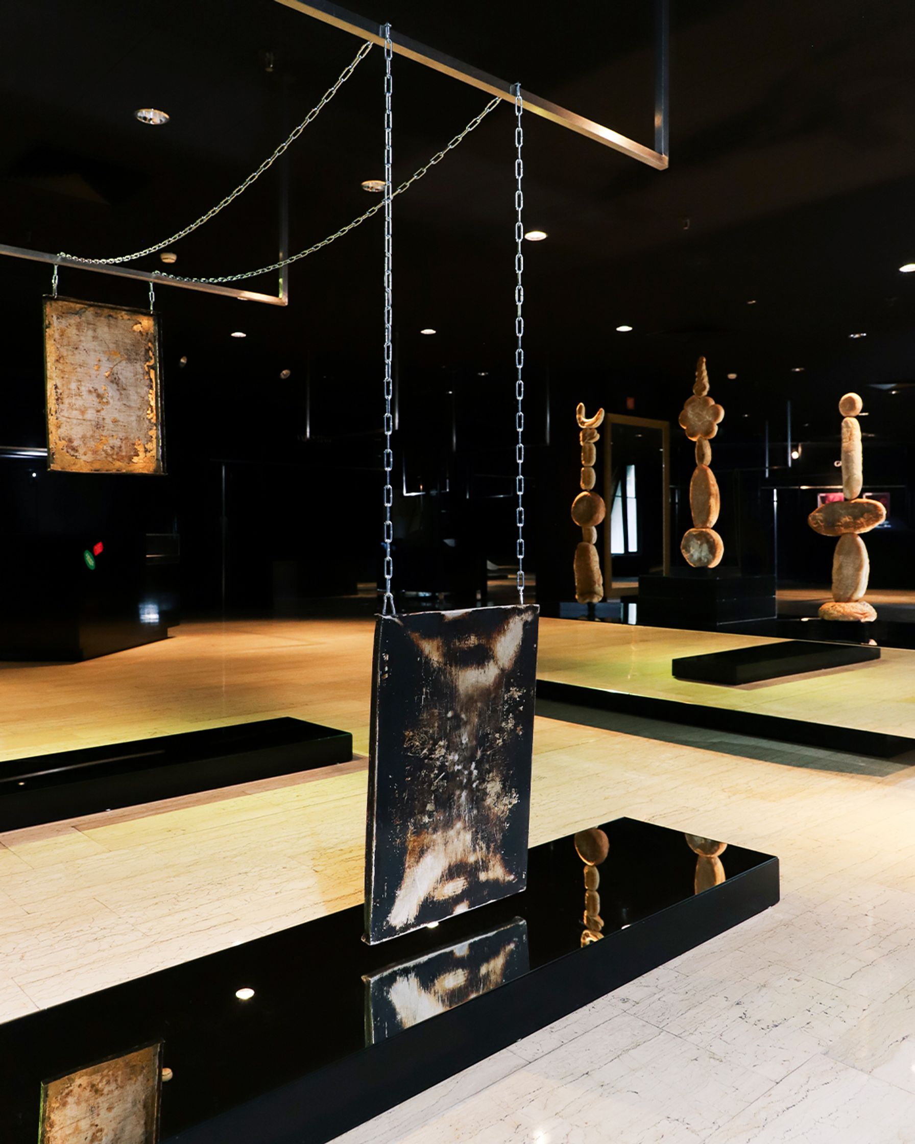 Rectangular panels featuring distressed black and white textures hang from chains over a shiny black reflective surface on a light-colored floor, with other textured sculptures in the background.