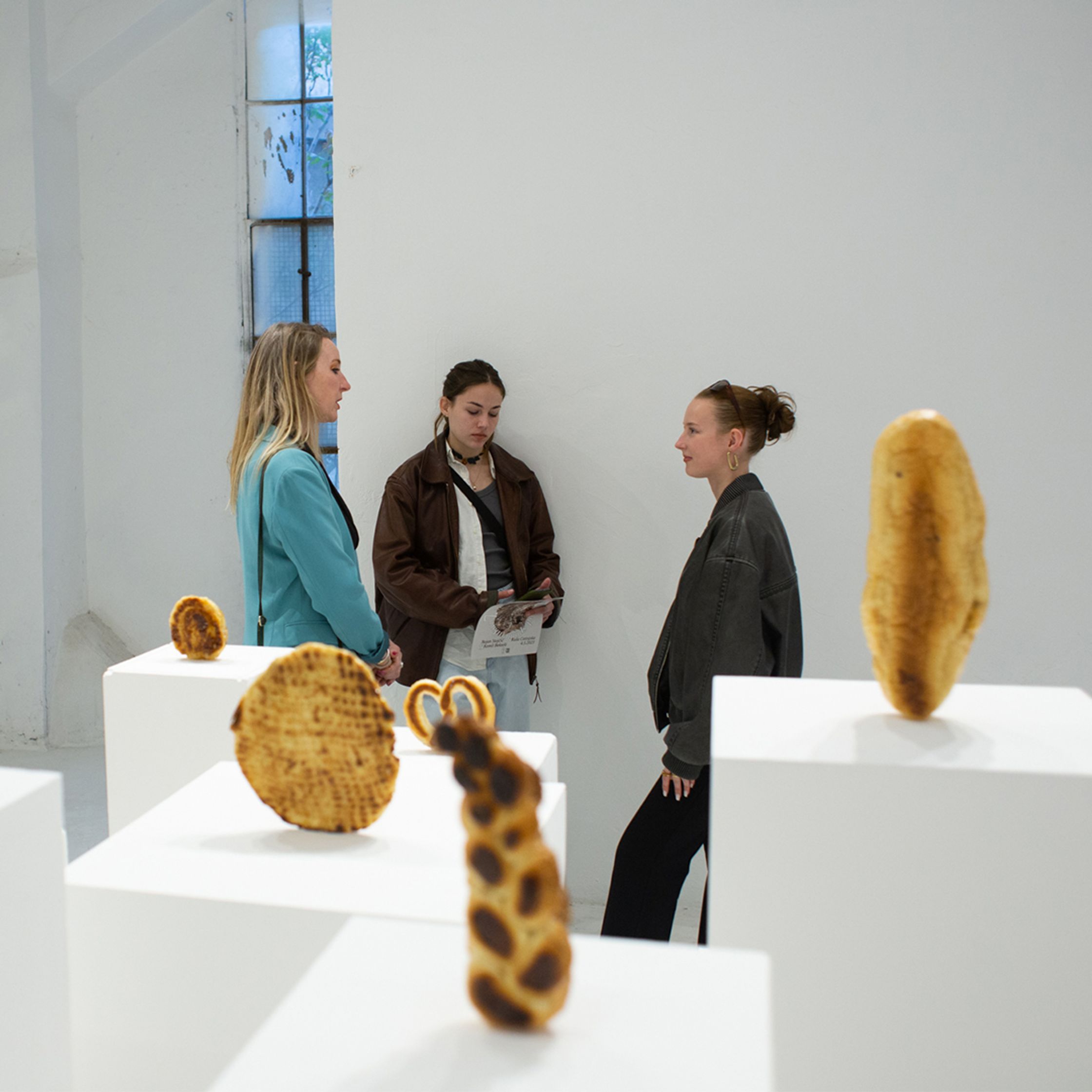 Three young women stand among an art installation of bread-like sculptures displayed on white pedestals in a gallery.