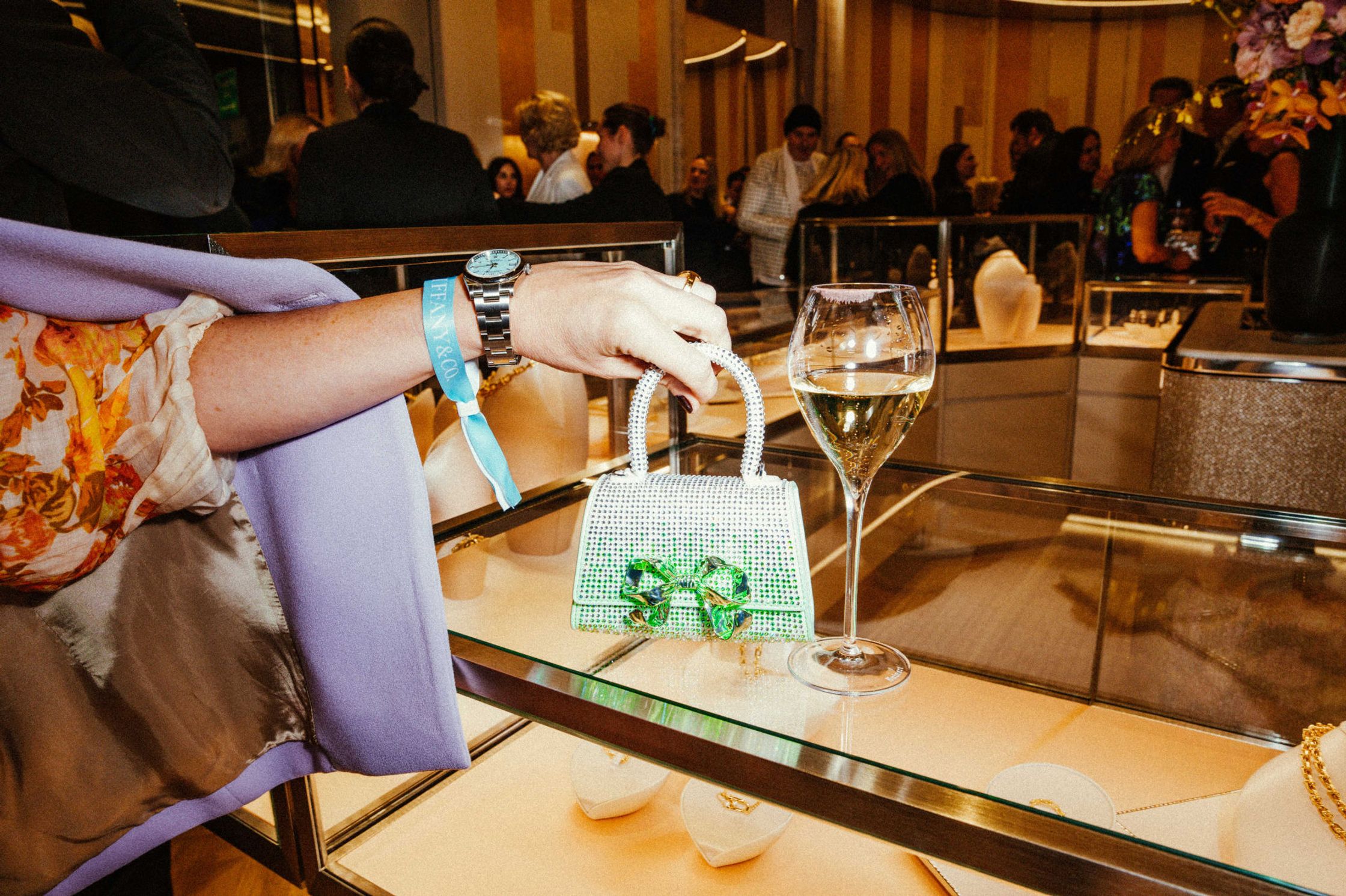 Frau mit Tiffany-Armband hält glitzerbesetzte Mini-Handtasche mit grüner Schleife auf einer Schmuckvitrine beim Tiffany & Co. Event Berlin – Champagnerglas daneben