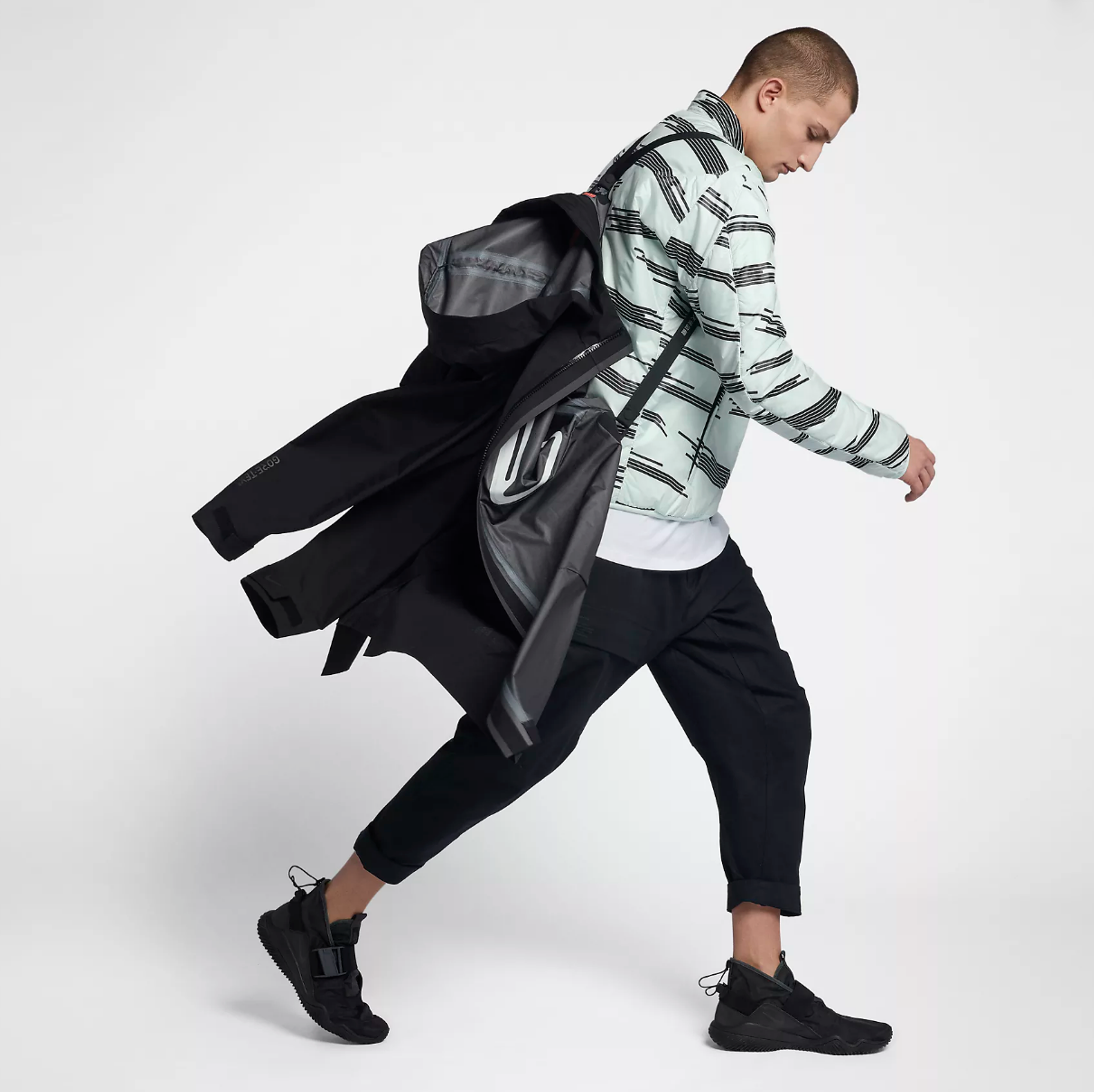 a man in a black jacket and black pants is walking on a white background nike