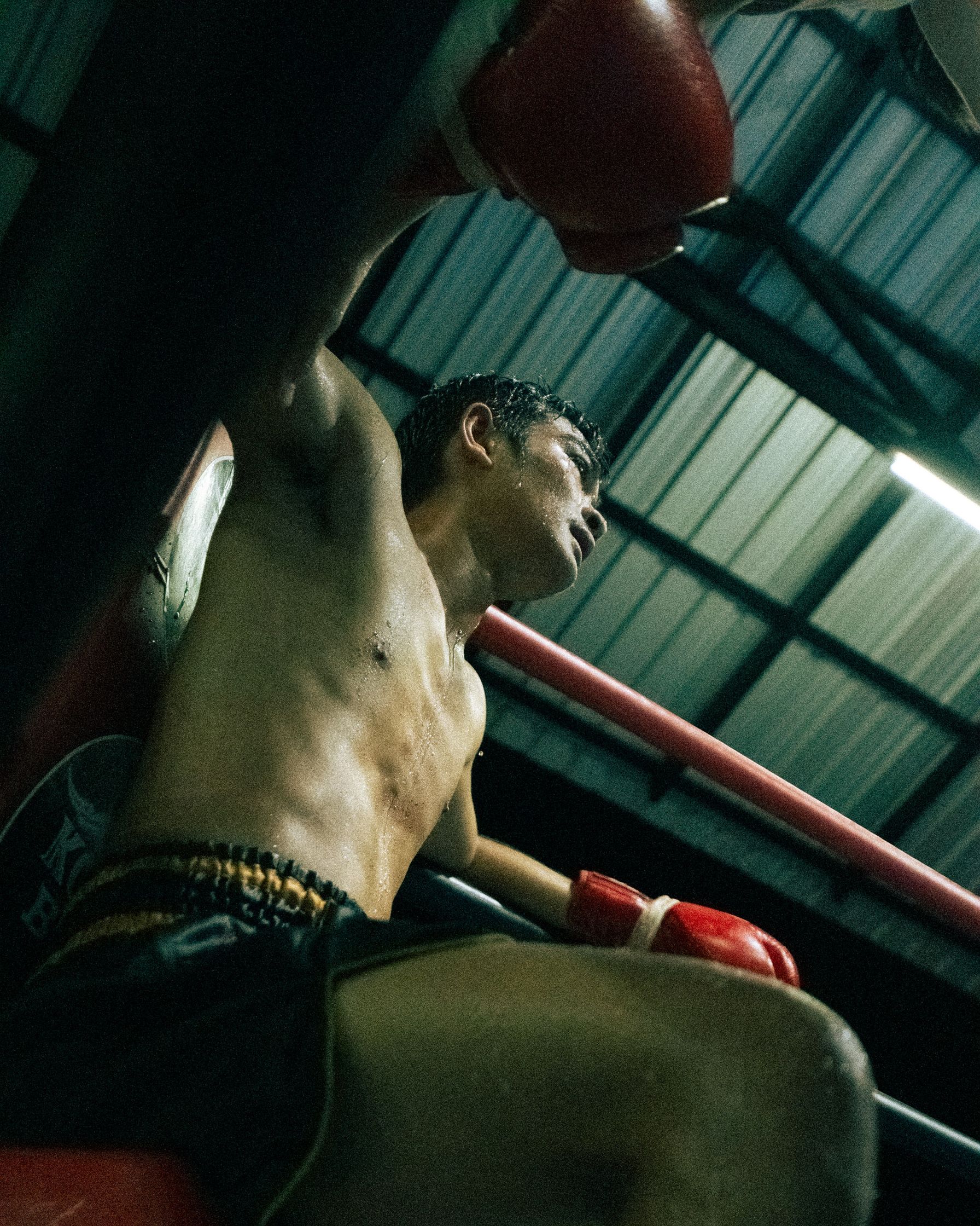 A raw moment of stillness: A Muay Thai fighter in his corner during the break, catching his breath amidst the heat and intensity of the stadium.