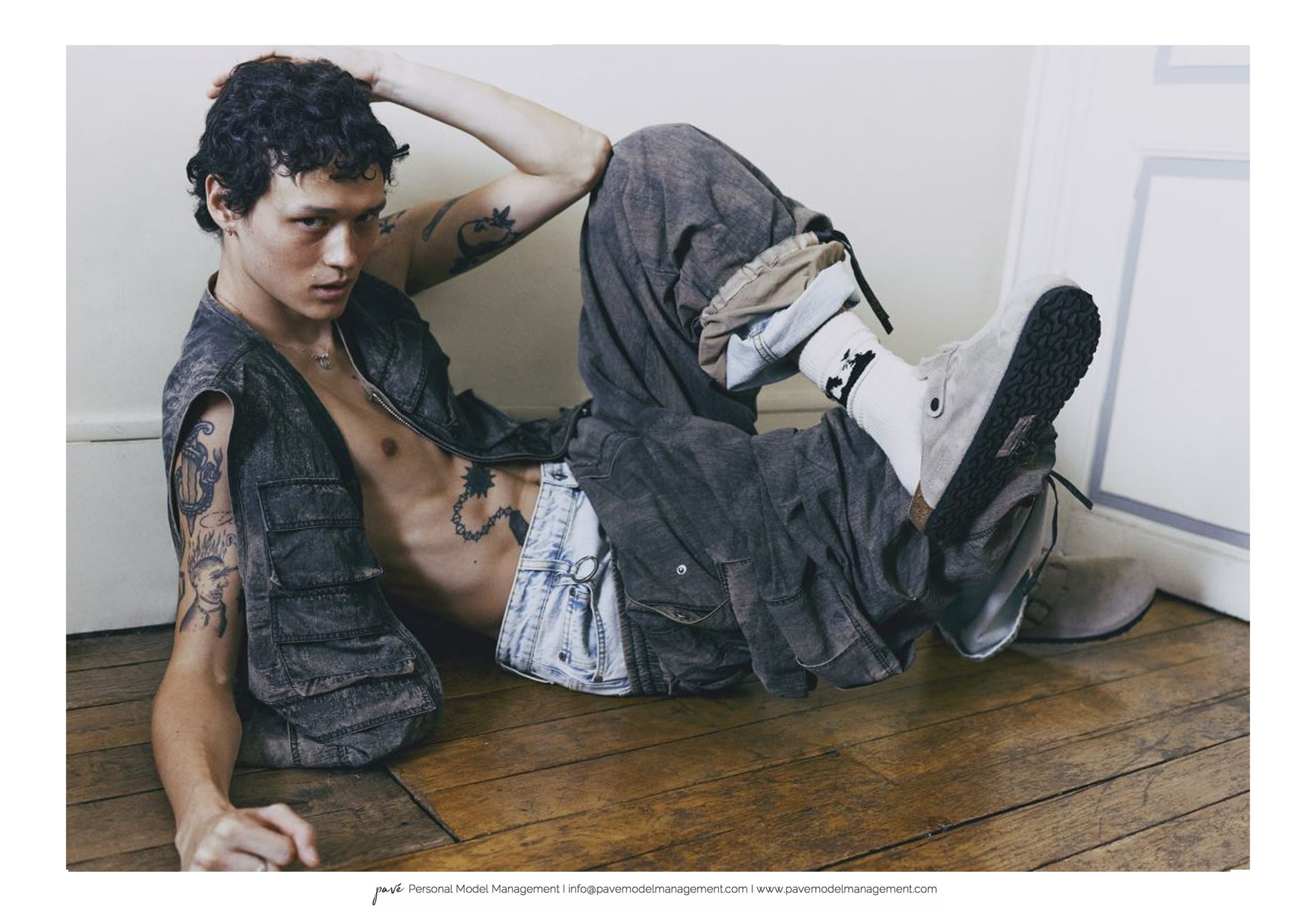 Fashion model with tattoos and curly hair posing in a vest, jeans, and loose pants.