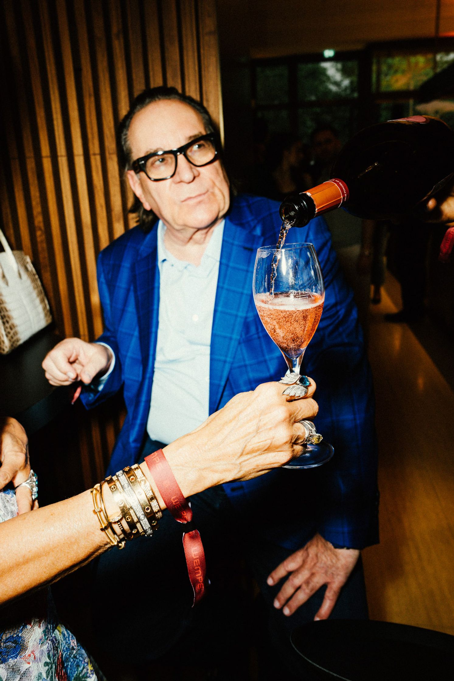 Stilvoll gekleideter älterer Herr im blauen Karojackett beim Rolex Boutique Event Berlin: Rosé-Champagner wird in sein Glas eingeschenkt, goldene Armbänder und Ring im Vordergrund