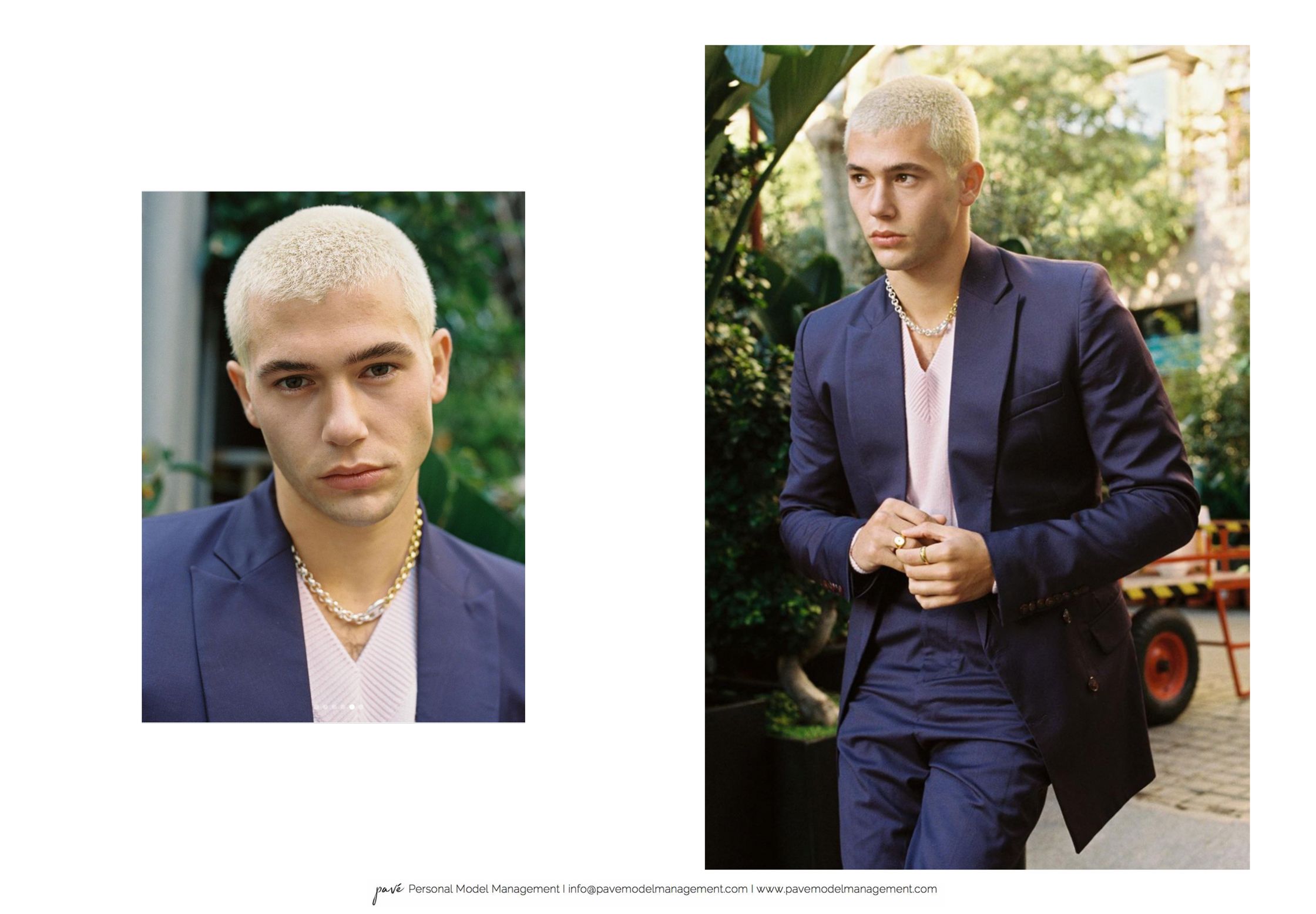 A light-skinned man with blond buzzcut hair wears a navy blue suit jacket over a pink sweater, with a chunky silver chain necklace, two silver rings on his left hand and one gold ring on his right hand, outdoors with green foliage in the background.