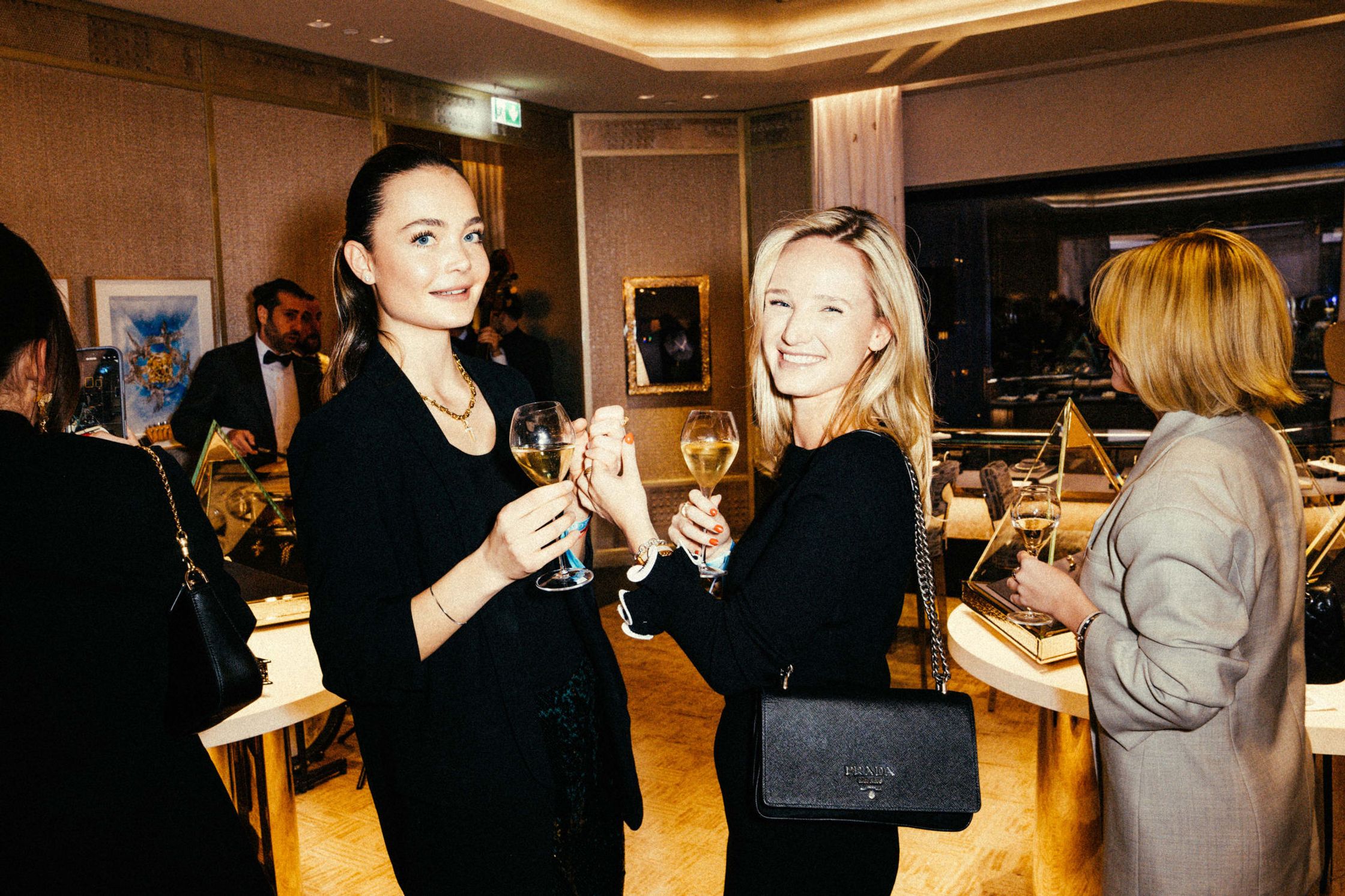 Zwei Frauen in Schwarz stoßen mit Champagner an beim Tiffany & Co. Event Berlin – eine trägt Prada-Tasche, Schmuckvitrinen mit goldenen Pyramiden im Hintergrund