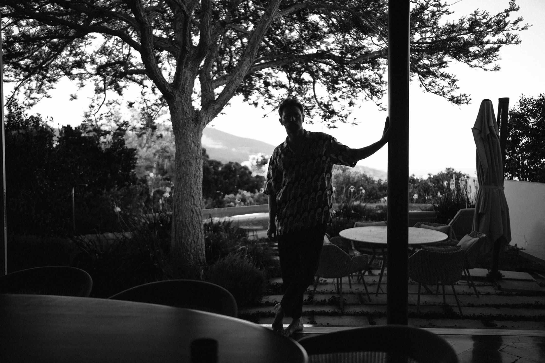 Künstler Paul Schrader als Silhouette in einer Glasschiebetür, Schwarz-Weiß-Aufnahme mit Baum und Bergpanorama im Gegenlicht, architektonisches Portrait Kapstadt 2024