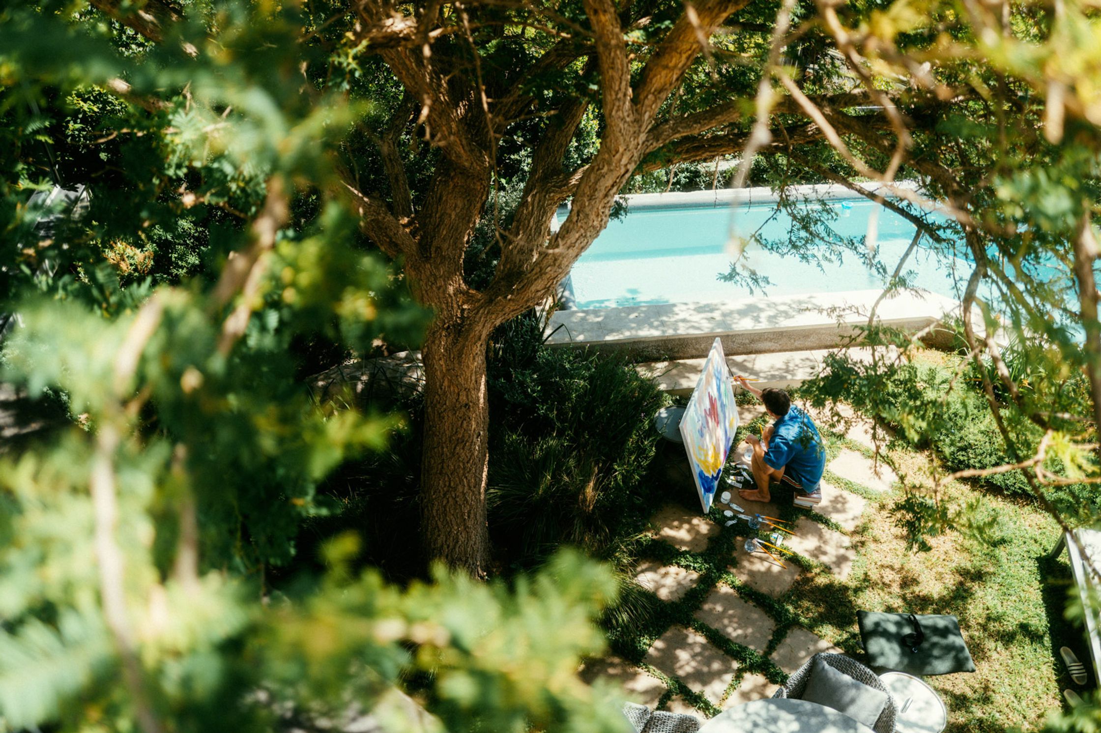 Draufsicht auf Künstler Paul Schrader beim Malen im Garten unter einem großen Baum, Schwimmbecken im Hintergrund, üppige Gartenlandschaft Kapstadt 2024