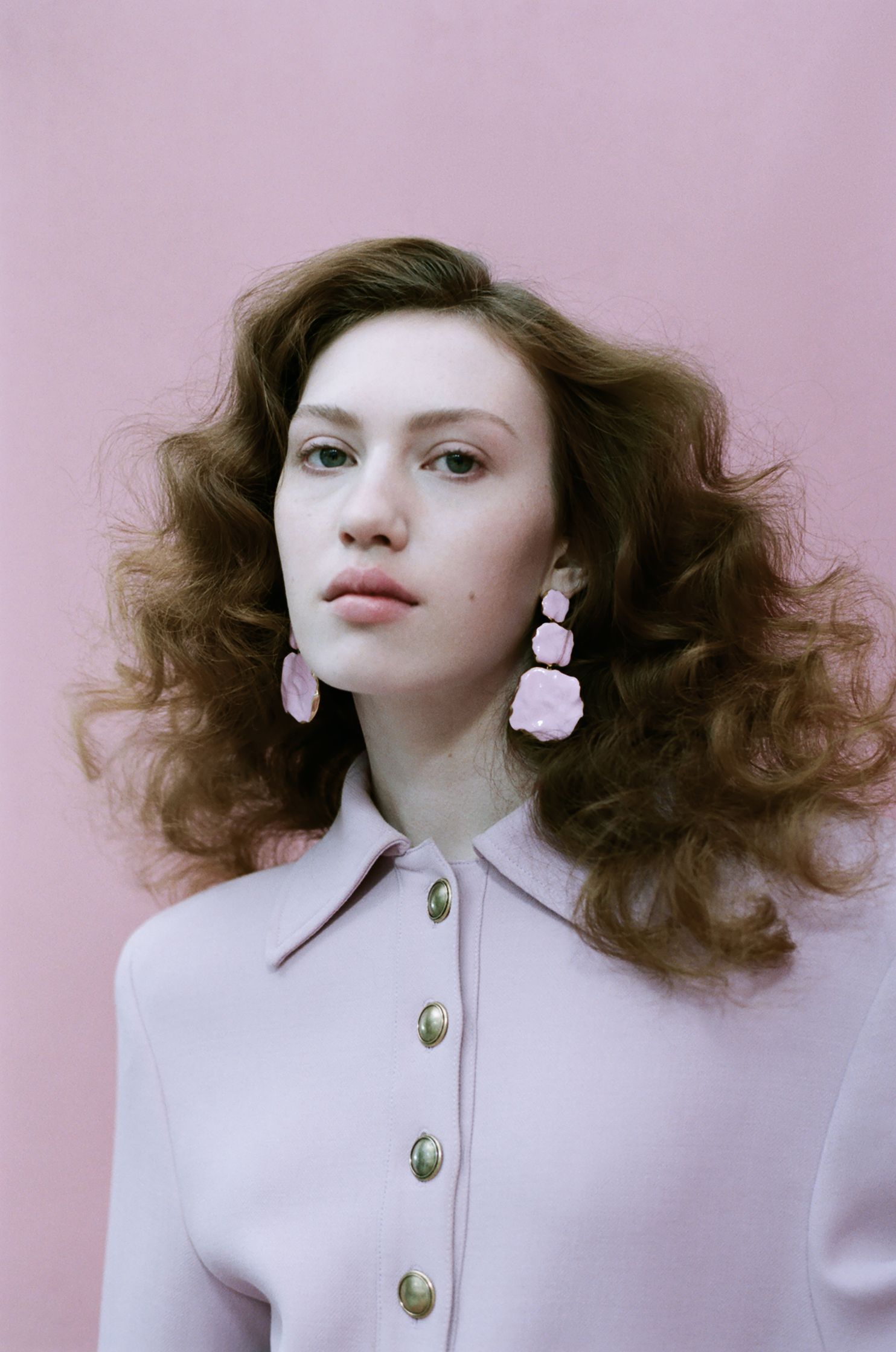 a woman with long curly hair and pink earrings posing against a pink wall