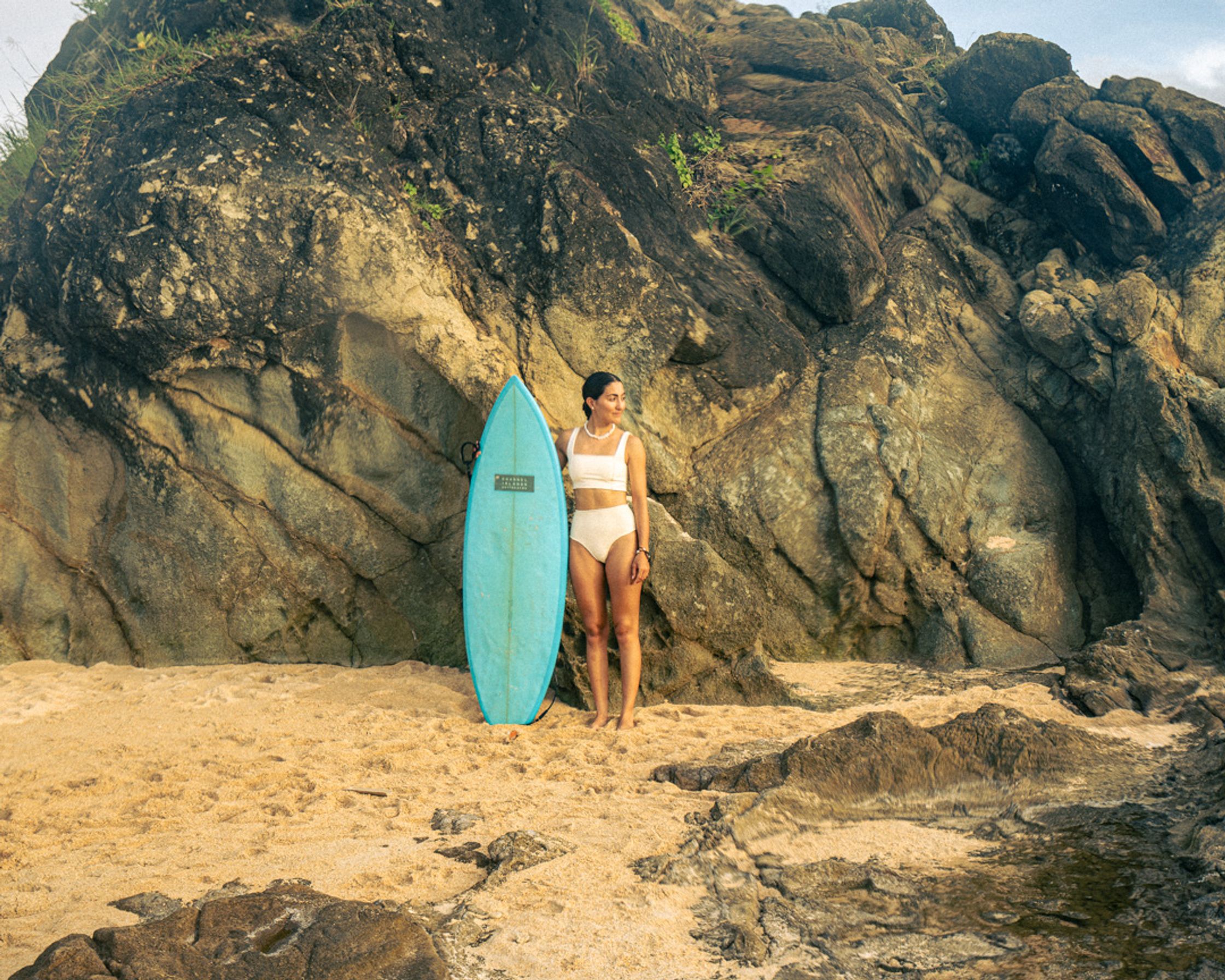 A surfer standing with her board on a rugged beach in South Lombok, in front of a large rock formation, thoughtfully looking out at the horizon.