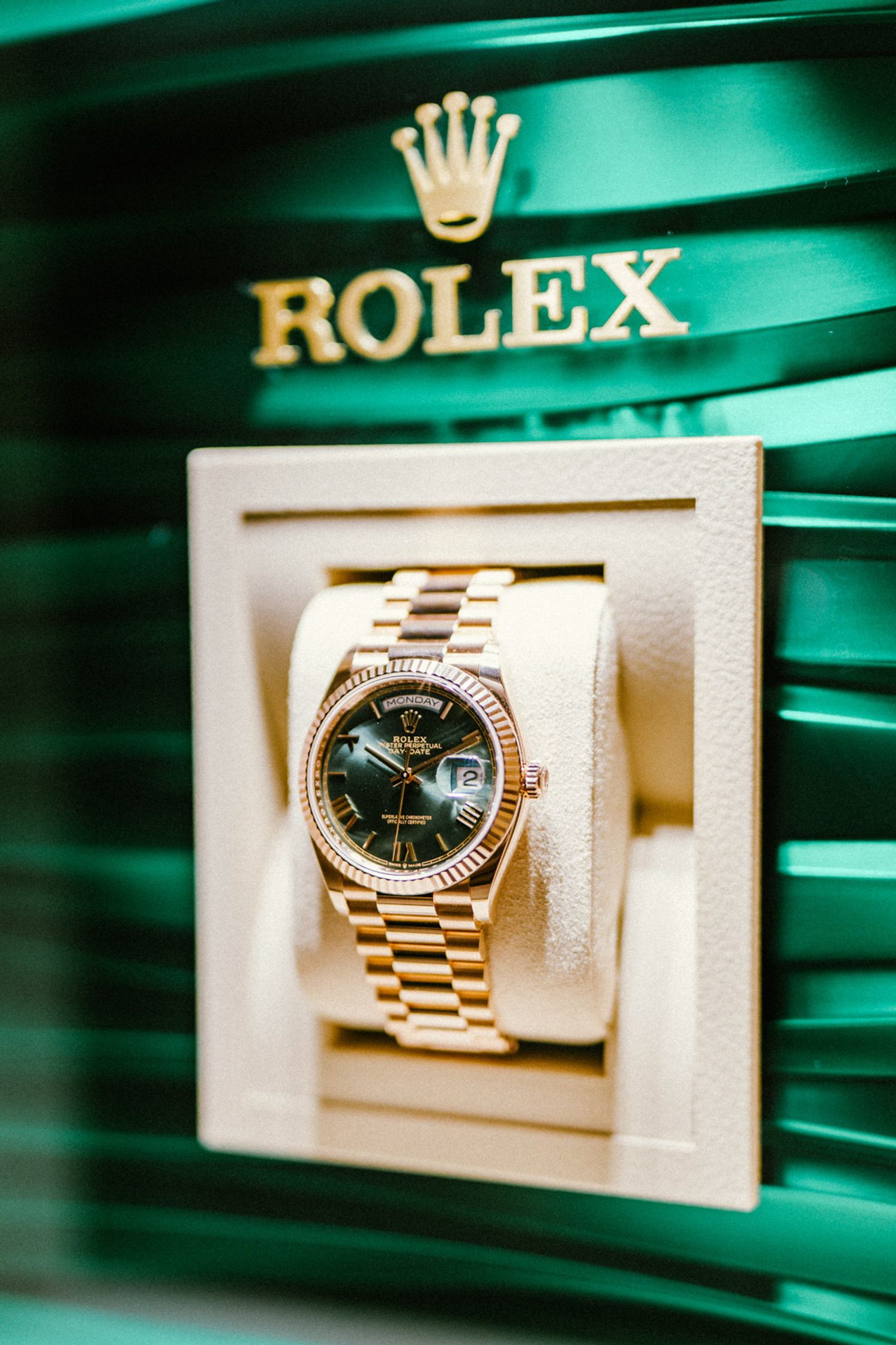 Rolex Day-Date in Gelbgold mit grünem Zifferblatt und geriefelter Lünette auf cremefarbenem Kissen vor ikonisch grünem Rolex-Schaufensterdisplay