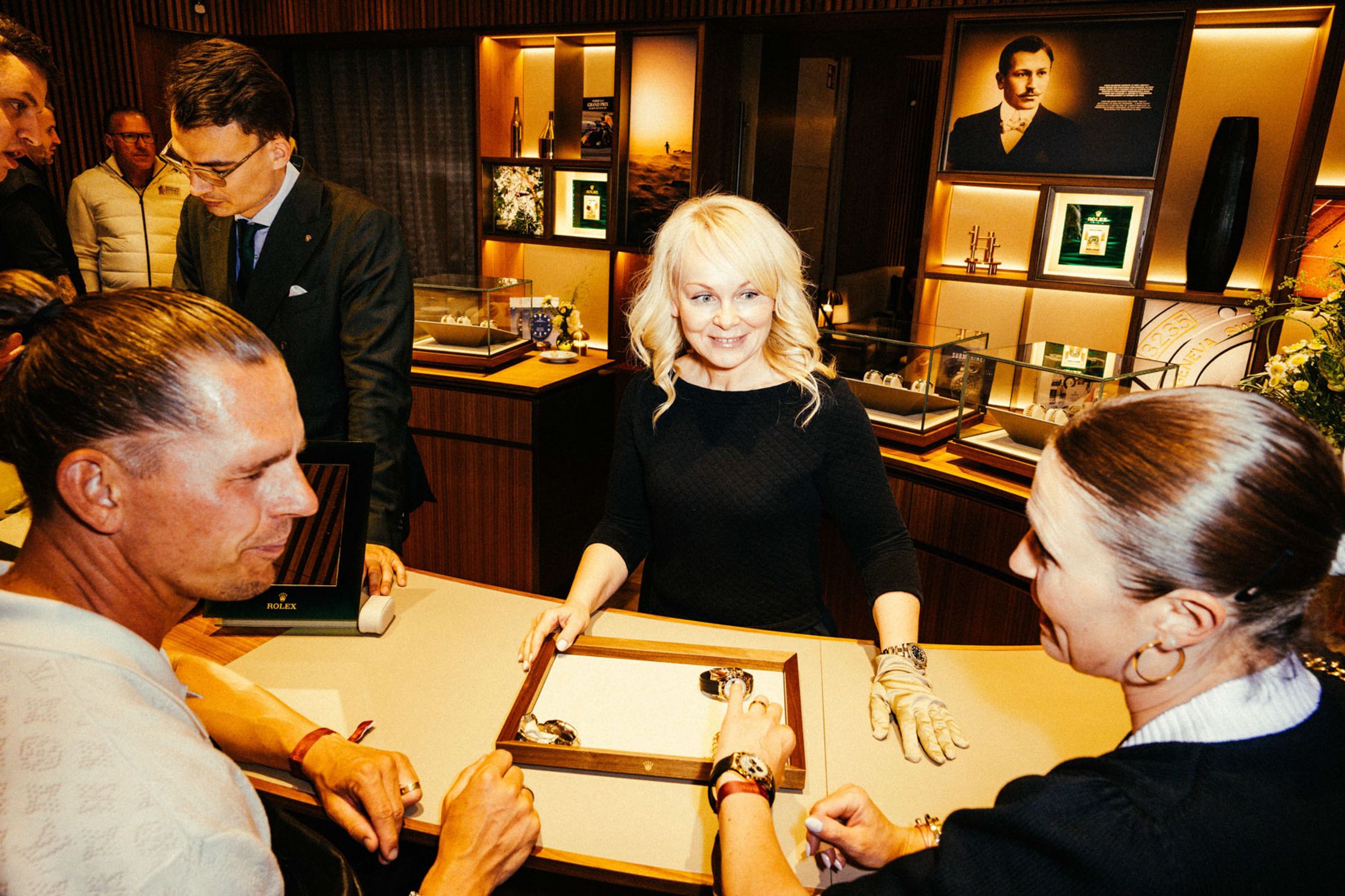 Beratungsszene in der Rolex Boutique Berlin: Blonde Frau im schwarzen Kleid und Kundenberaterin präsentieren einem Kunden eine Rolex-Uhr auf einem Tablett, elegantes Luxus-Uhrgeschäft Interieur