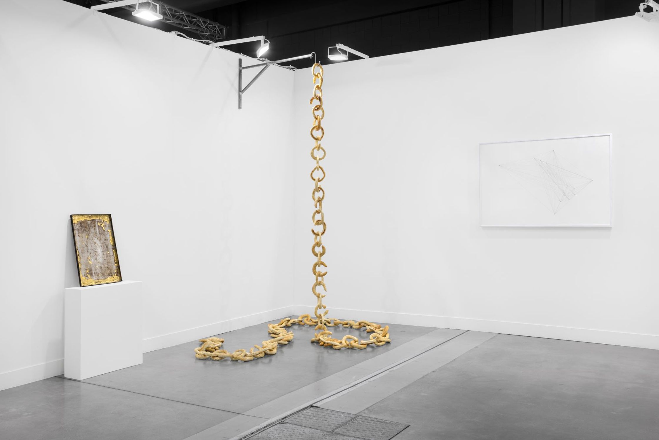 A beige chain hangs from the ceiling and coils on the floor, next to a framed artwork on a white pedestal and a framed sketch on the wall, all in a white gallery space.