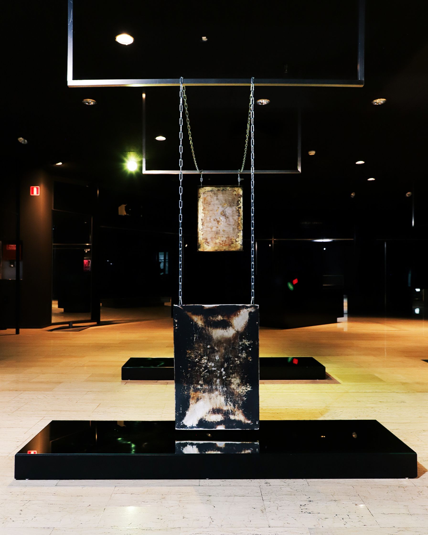 An art exhibition featuring two rectangular, distressed metal plates suspended by chains above a reflective black platform in a dimly lit room.
