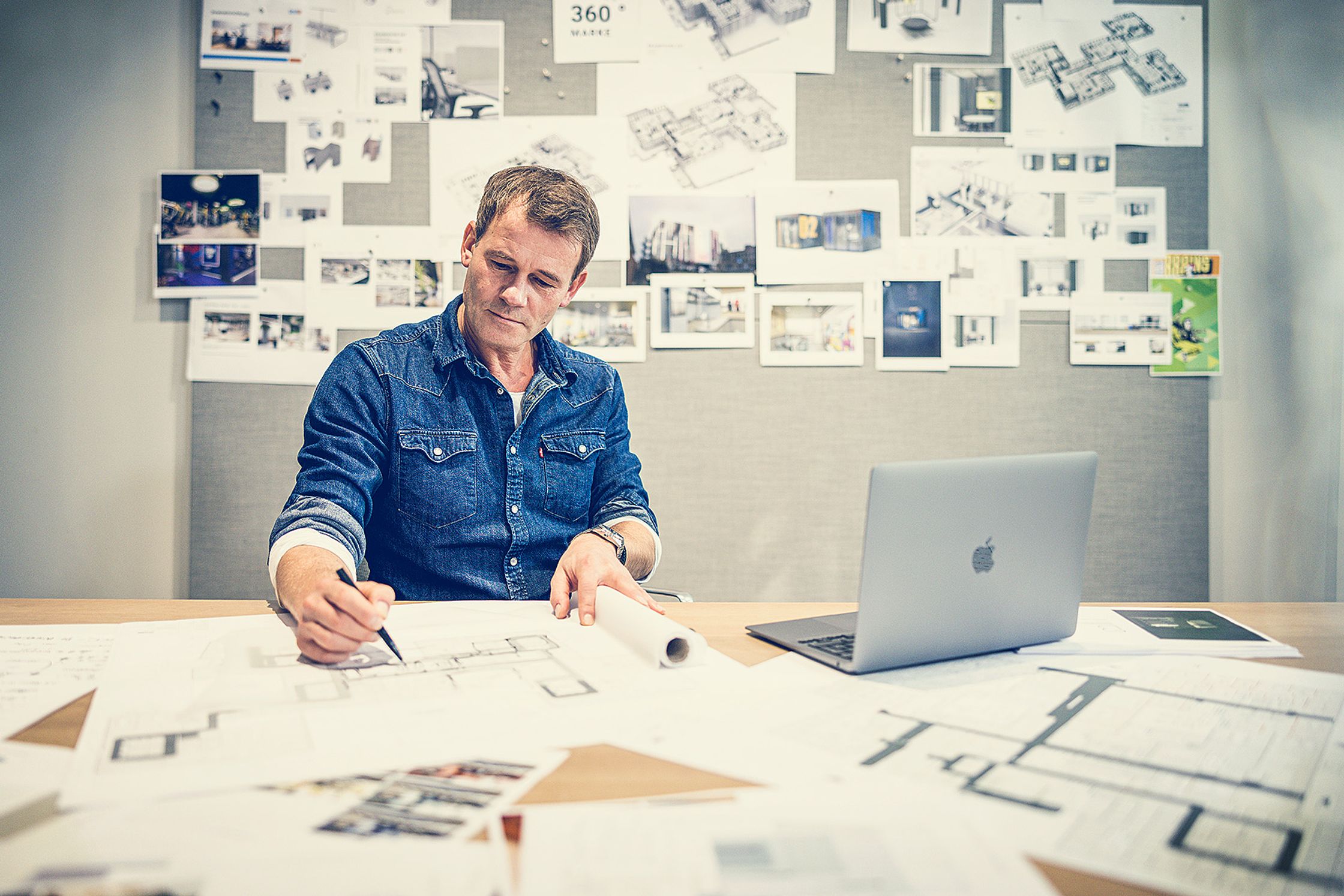 An architect in a denim shirt drawing blueprints at his desk with a laptop, in front of a mood board. corporate portrait photography