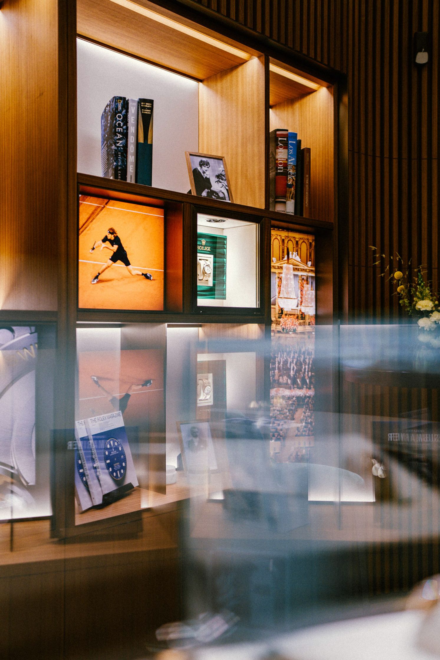 Beleuchtetes Holzregal in der Rolex Boutique Berlin mit gerahmten Bildern eines Tennisspielers, Rolex-Werbemotiven und Coffeetable-Books