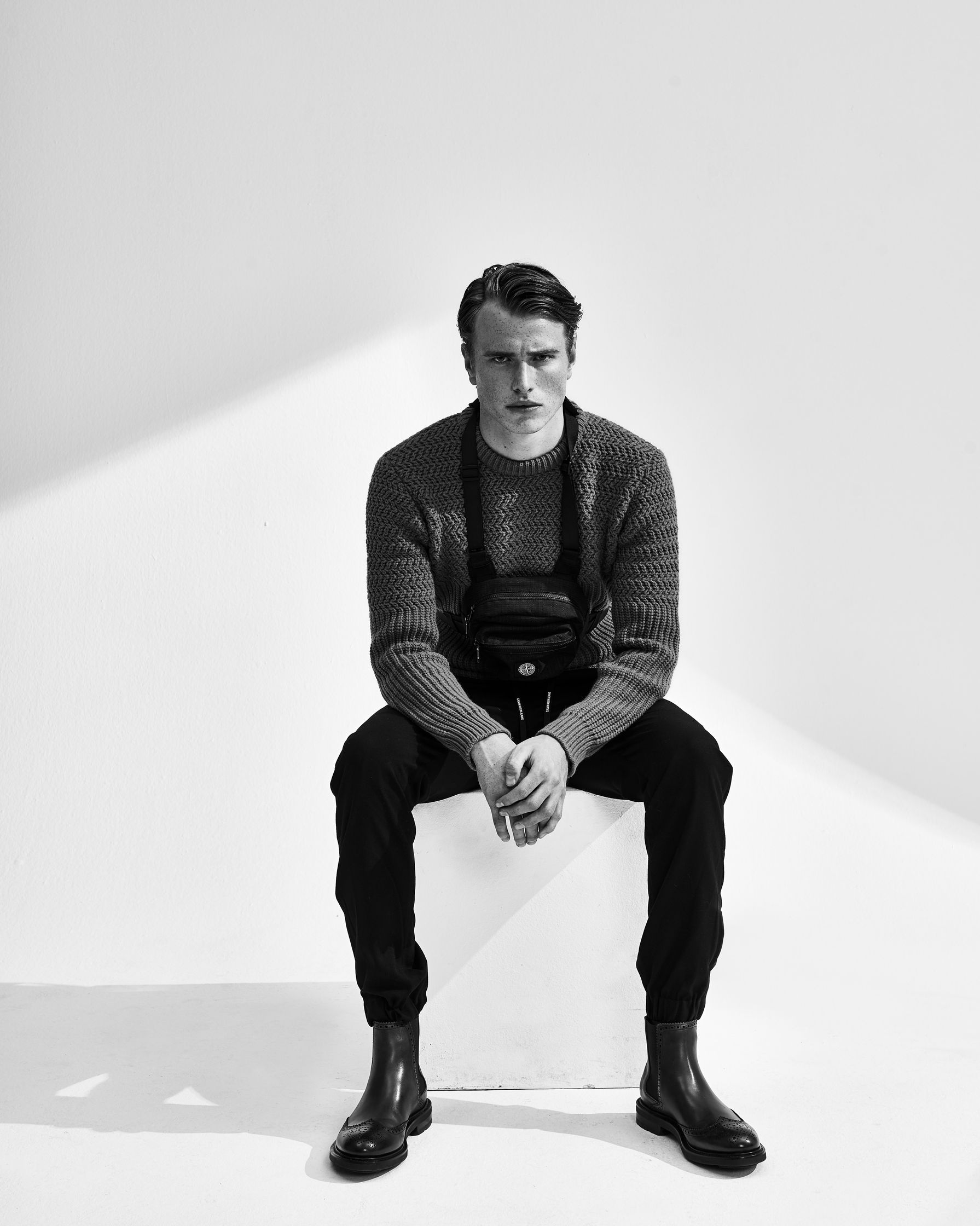 a man sitting on a white cube wearing a sweater and boots