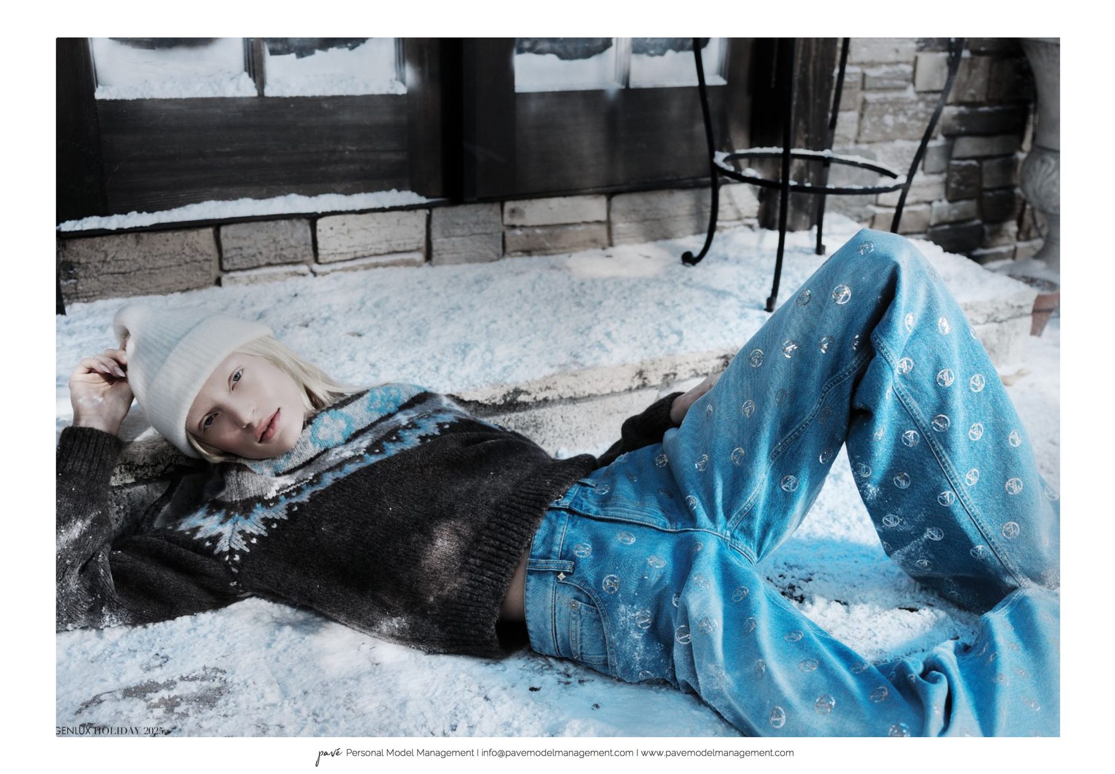 A model wearing a white beanie, black and blue knitted sweater, and peace sign jeans lies in the snow.