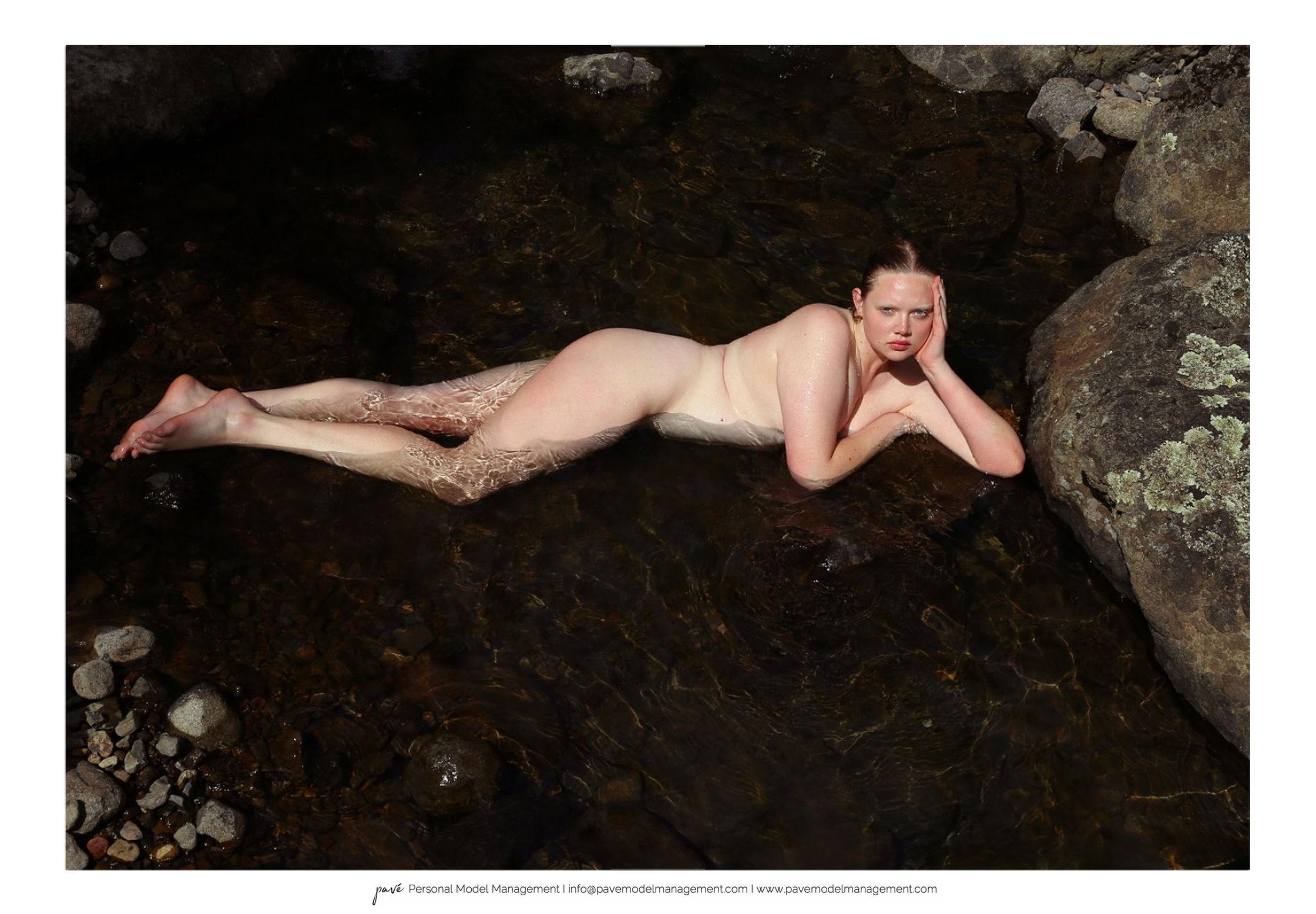 A pale woman lays nude in a dark, rocky stream, her light skin contrasting with the dark water.