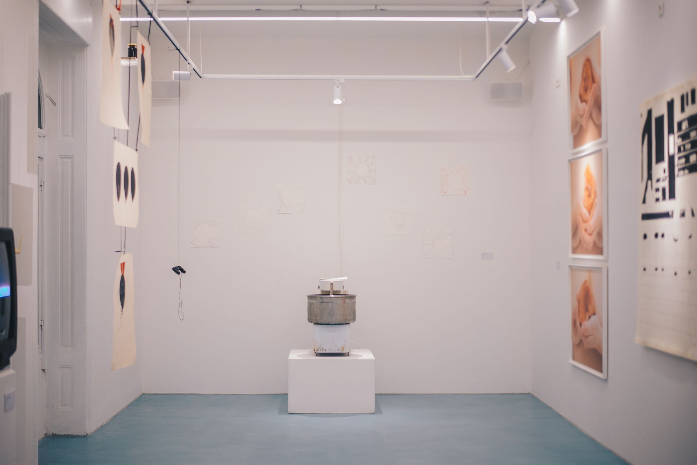 A brightly lit gallery space with a light blue floor, white walls displaying artwork, and a pedestal in the center.