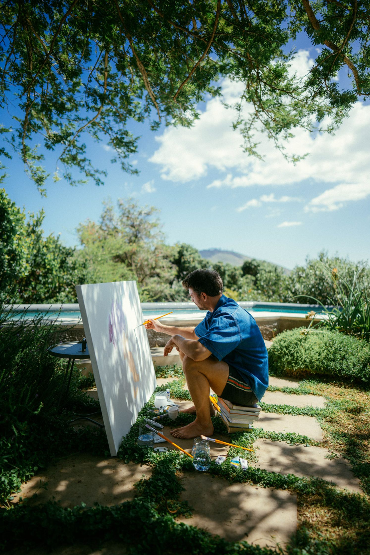 Maler Paul Schrader arbeitet konzentriert an einer Leinwand im Freien – hockend in einem schattigen Garten in Kapstadt, Pinsel in der Hand, mit Pool und Bergen im sonnigen Hintergrund