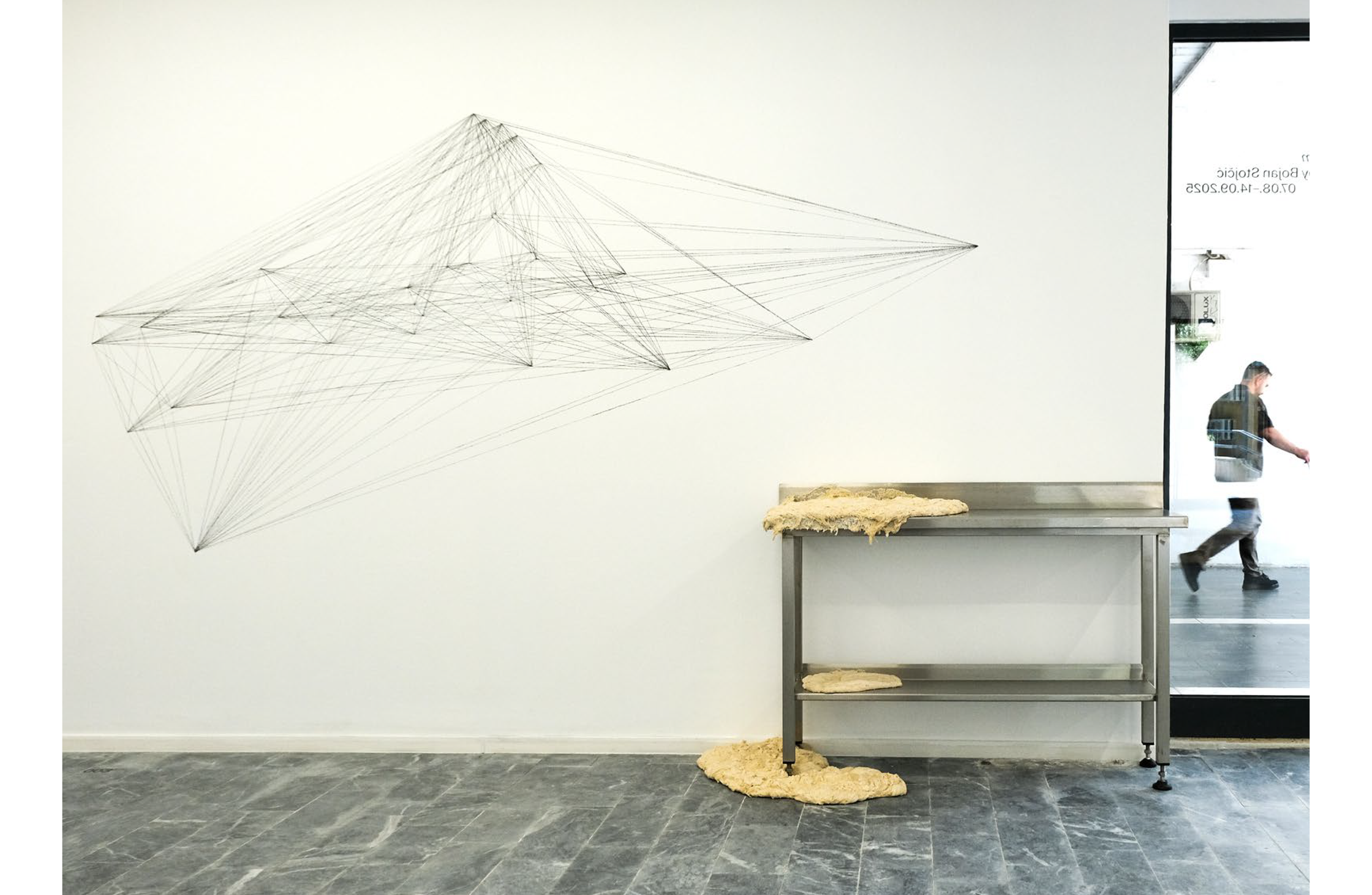 A white gallery wall featuring a geometric black string sculpture and a metal table with a beige, textured substance spilling from it, next to a reflective glass door showing a man walking.