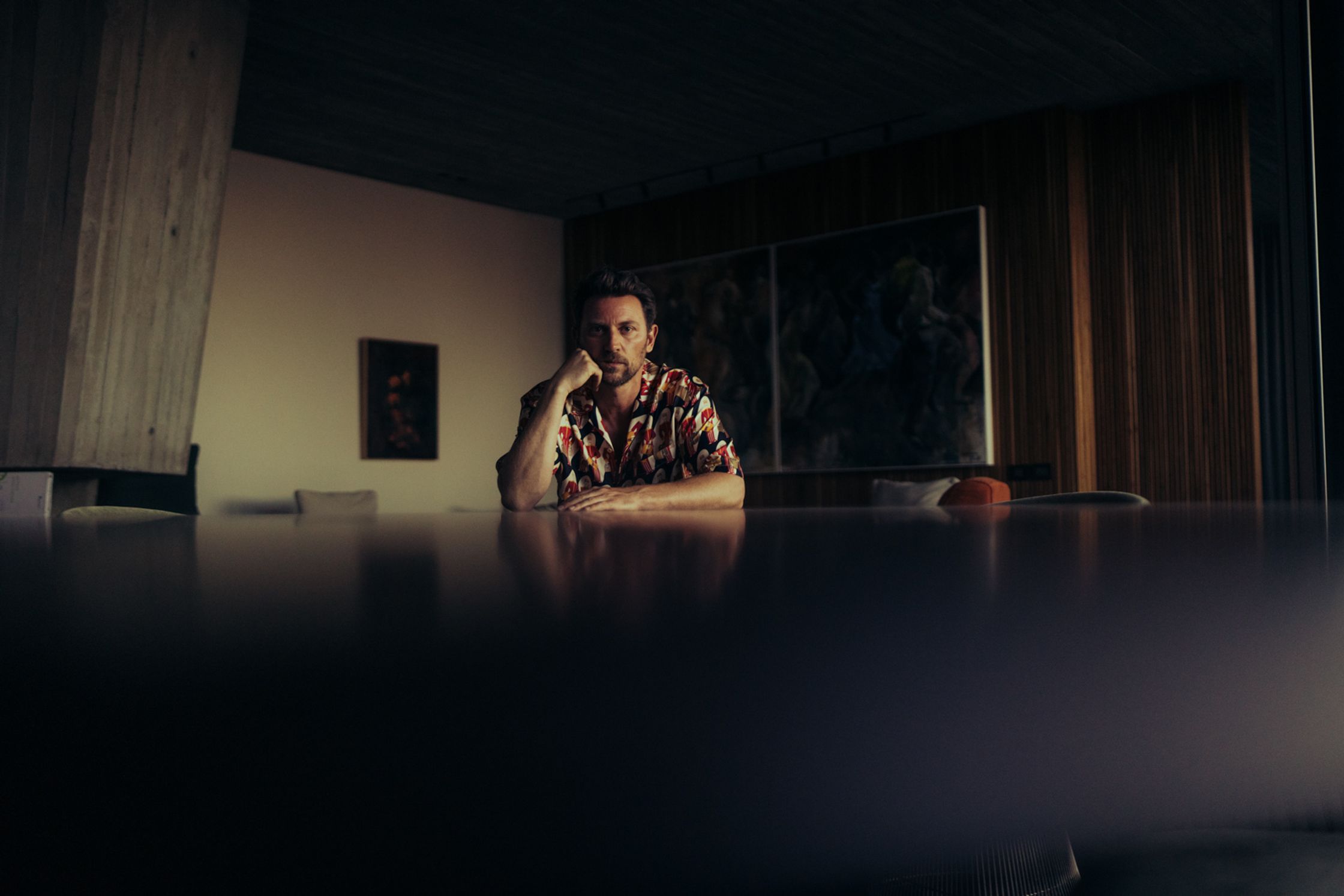Künstler Paul Schrader sitzt nachdenklich an einem großen Tisch in dunkelm stimmungsvollem Interieur mit Gemälde an der Wand, florales Hemd, Portrait-Fotografie Kapstadt 2024