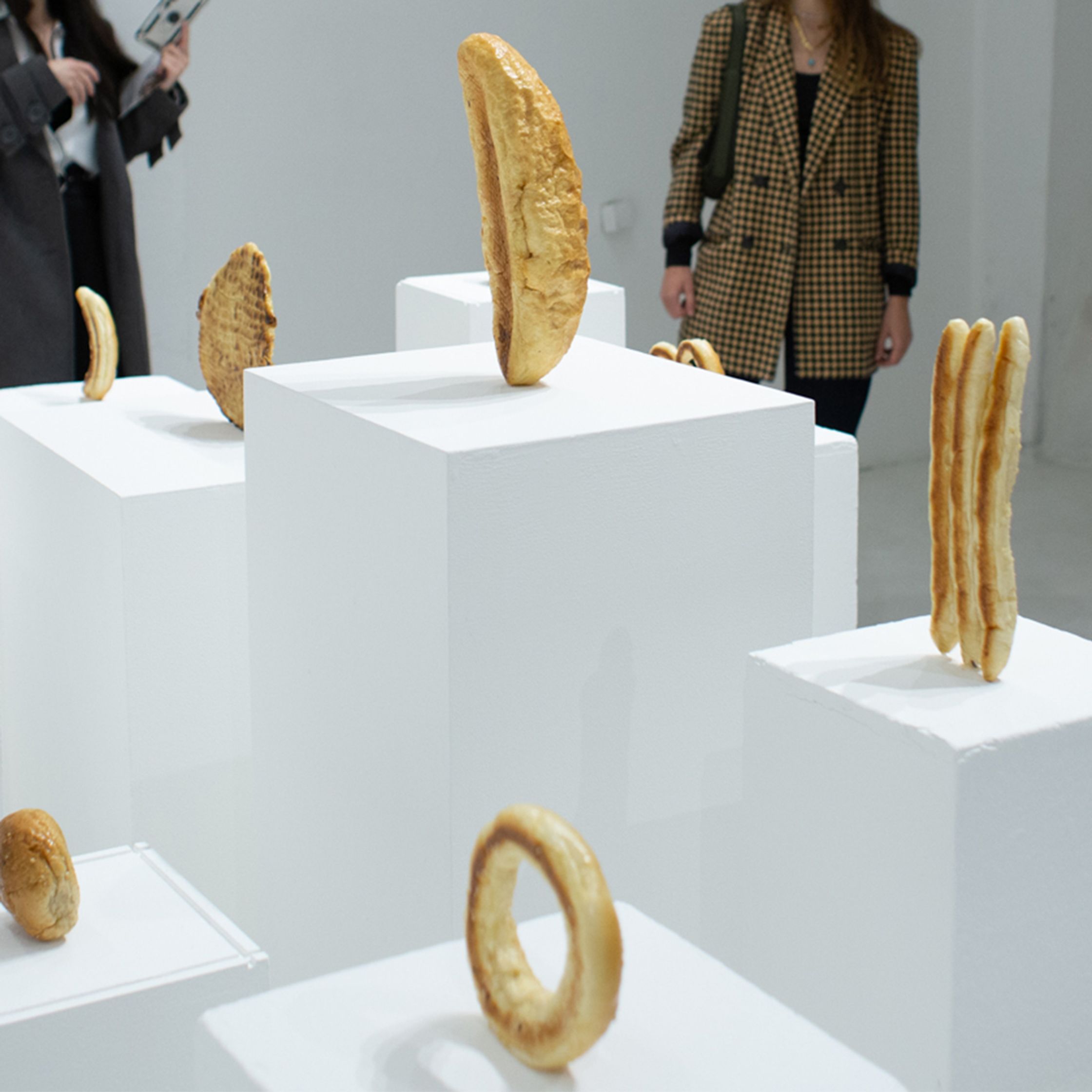 Various bread-like sculptures are displayed on white pedestals in a gallery with two people partially visible in the background.
