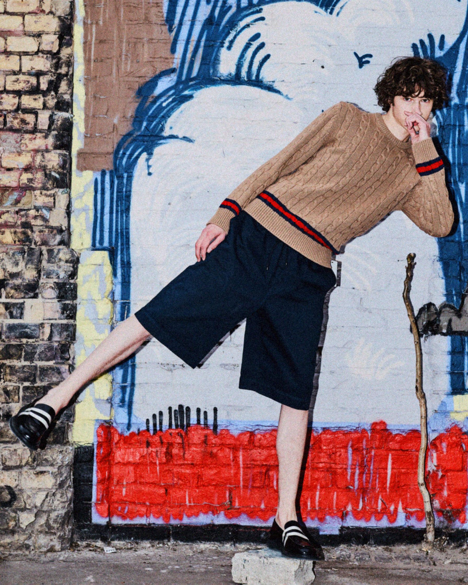 a man leaning against a wall in a sweater and shorts