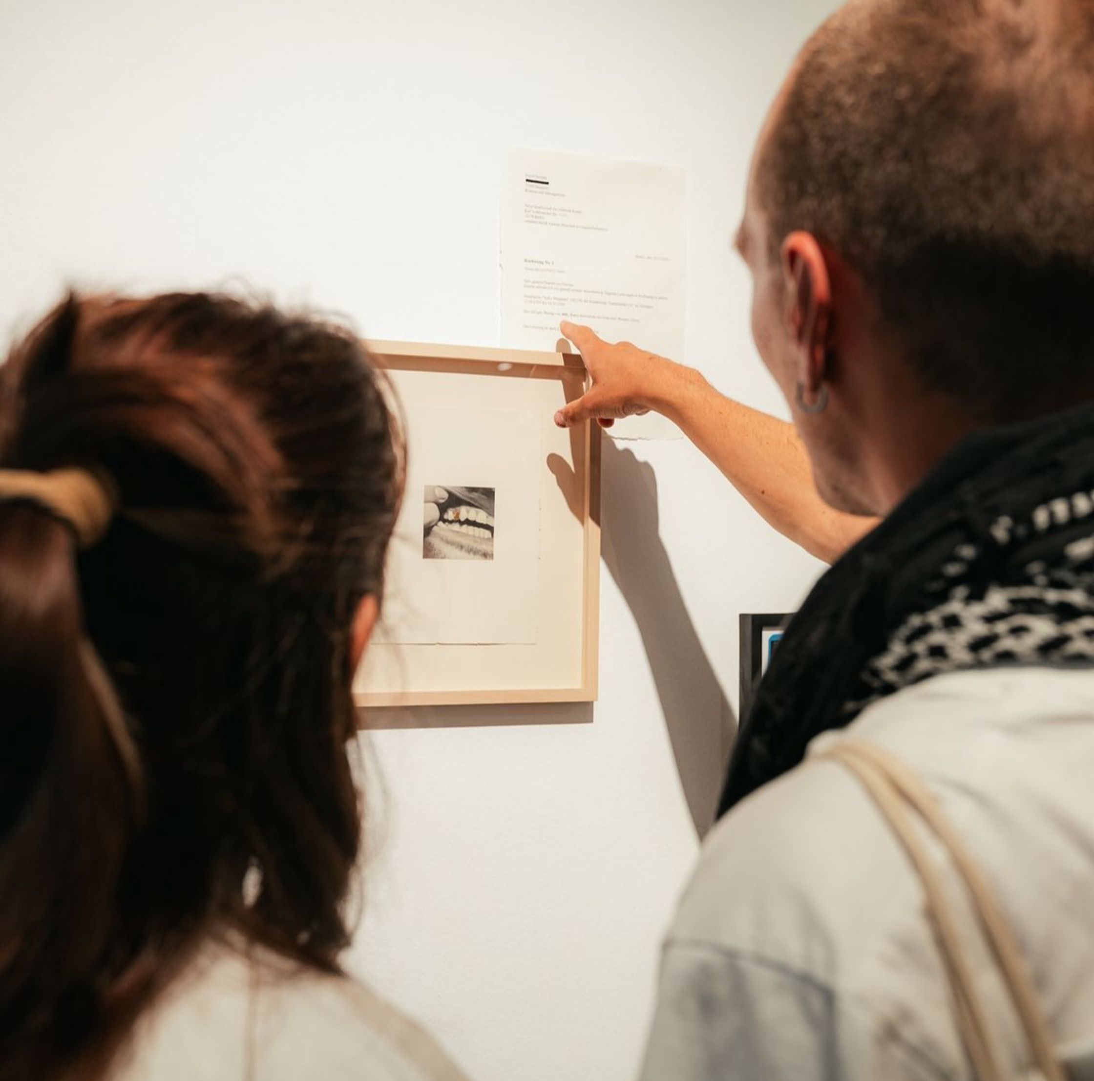 Two people looking at a framed image of teeth with gold caps and a document on a white wall.