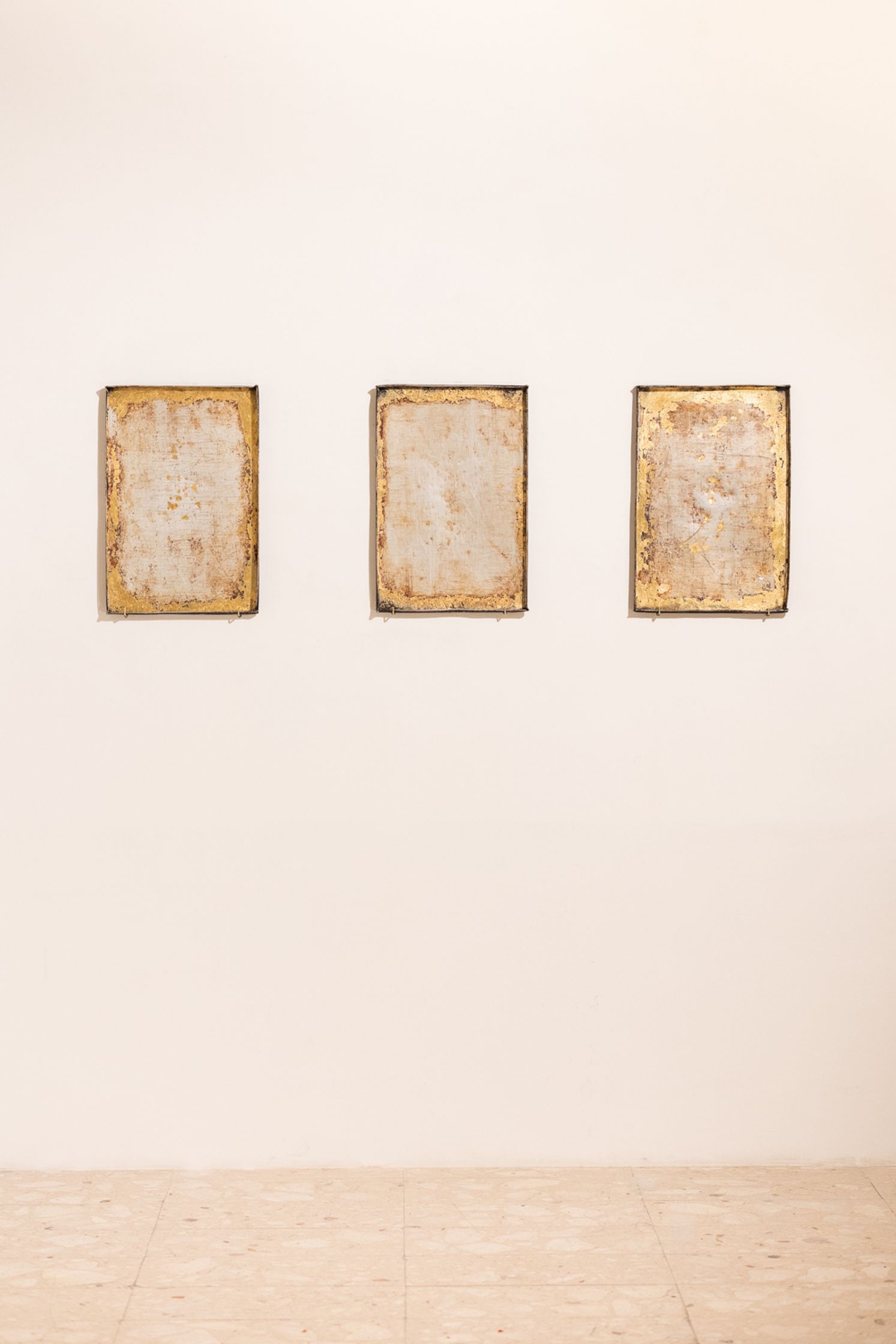 Three rectangular artworks with distressed gold frames and muted, neutral-toned centers hang on a plain light wall above a tiled floor.