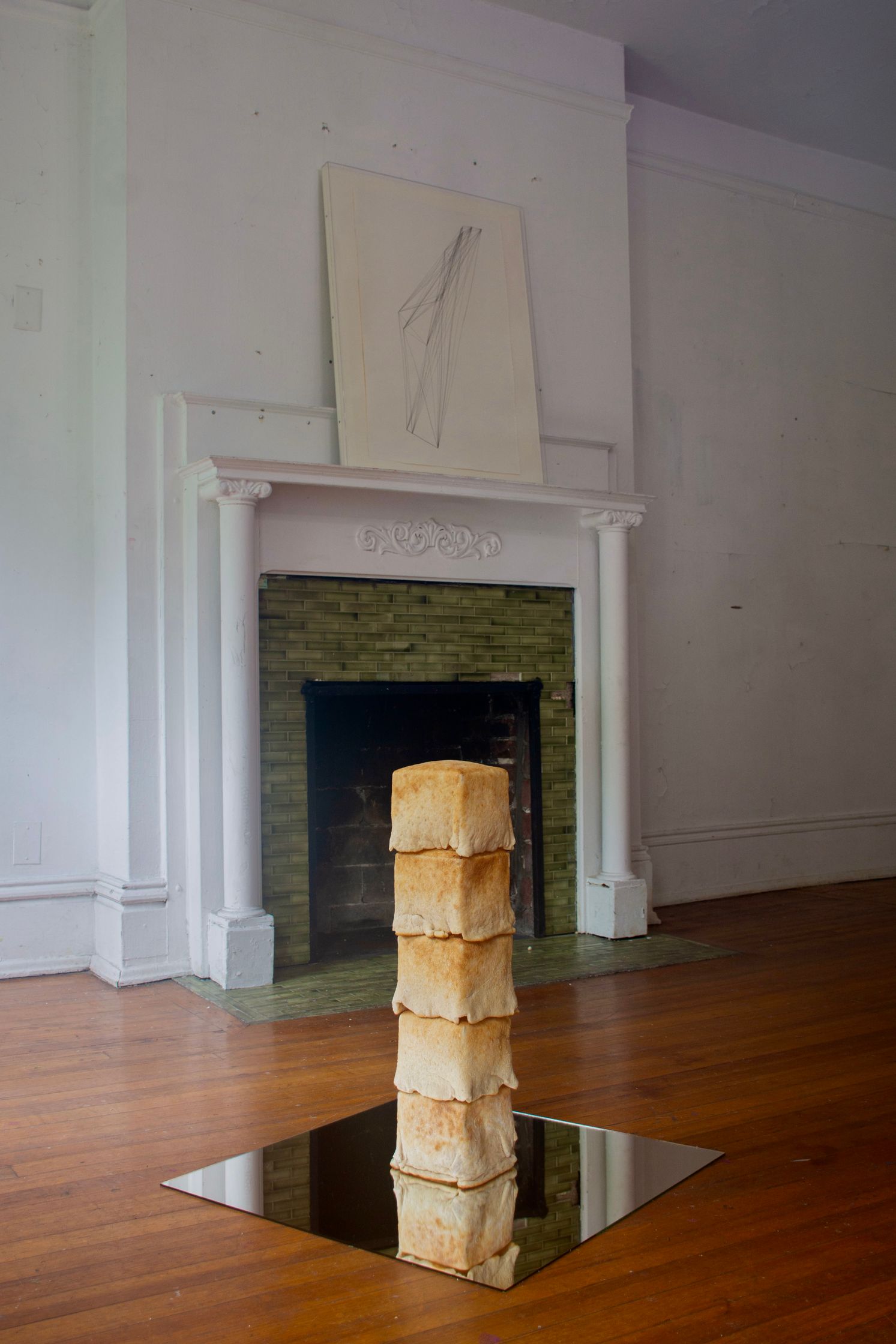 A tower of four square bread loaves on a mirrored surface, in front of a white fireplace with a drawing on the mantelpiece, in a room with hardwood floors.