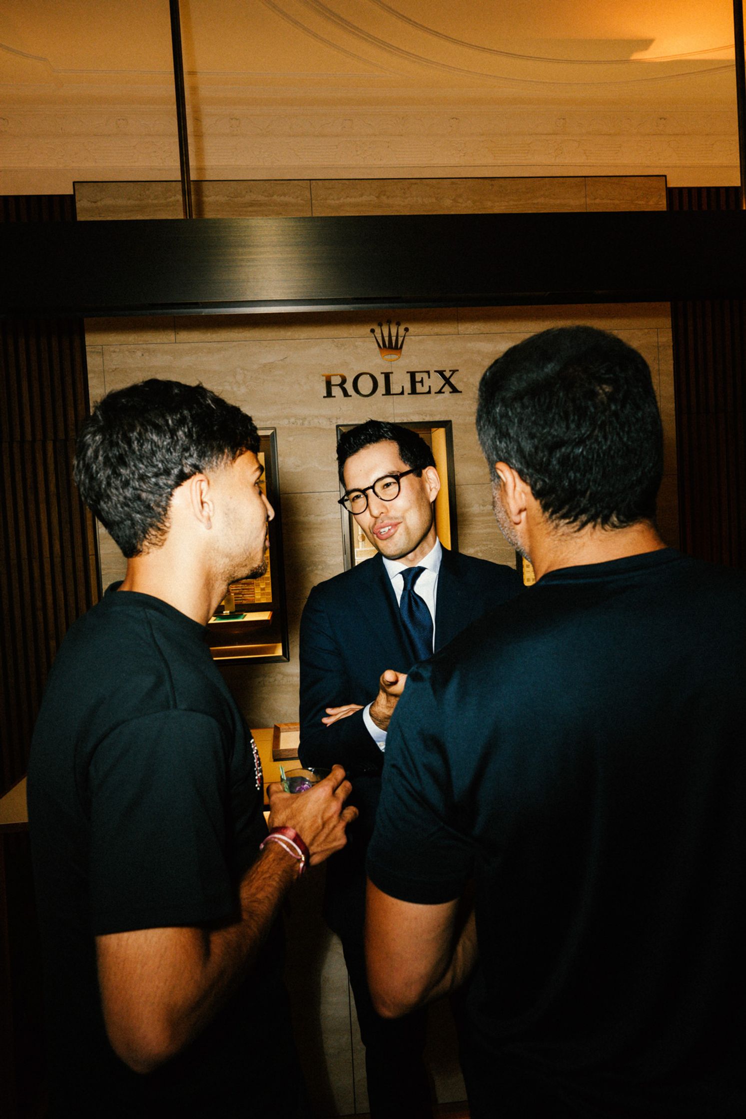 Drei Männer im Gespräch beim Rolex Boutique Berlin Event: Eleganter Herr im dunklen Anzug mit Brille berät zwei Gäste in schwarzen T-Shirts, Rolex-Logo auf Holzpaneel im Hintergrund