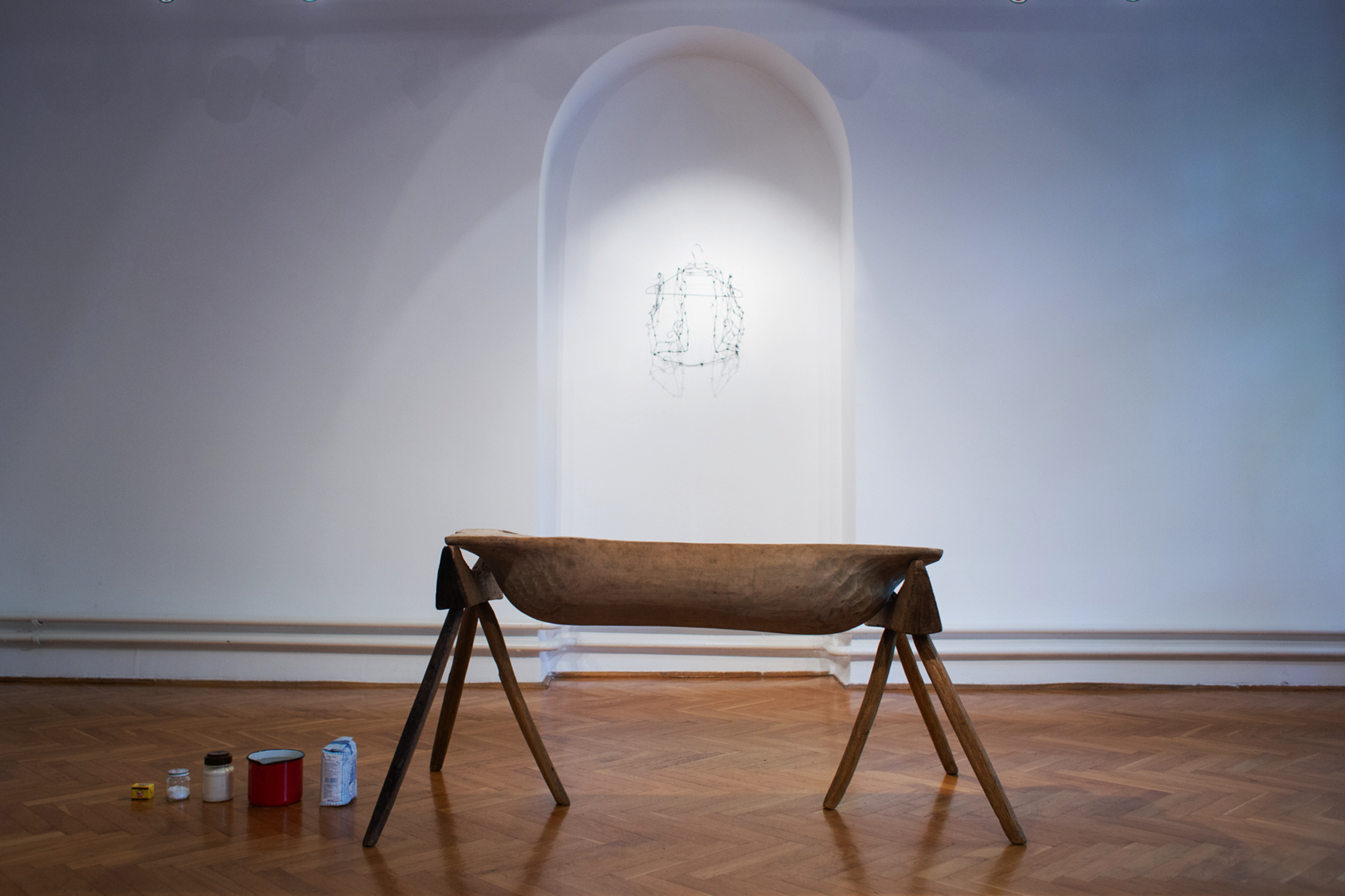 A wooden trough on tripod legs with cooking supplies and a wire sculpture in an arched alcove.