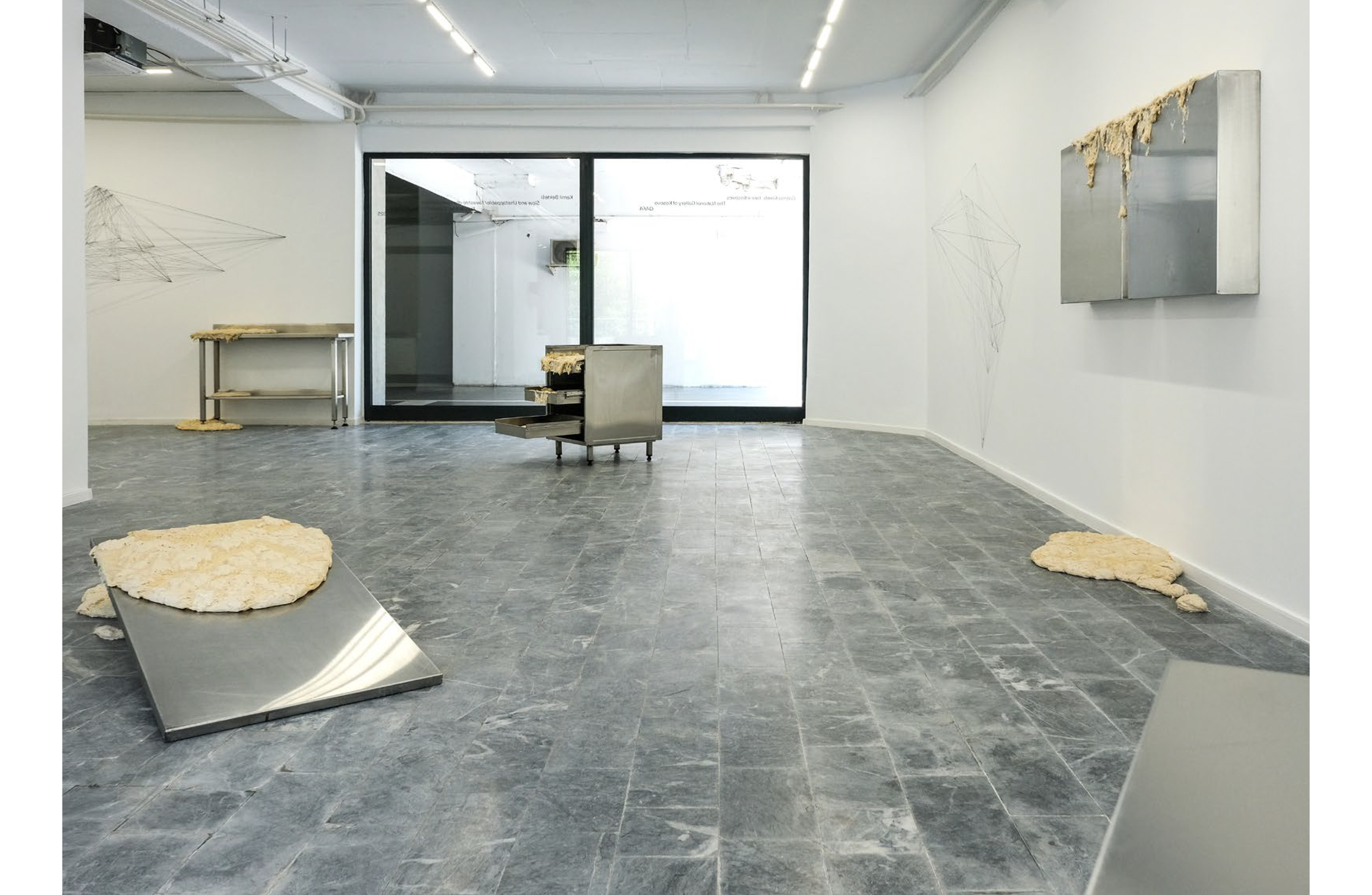 An art exhibit featuring raw dough on metal surfaces and on a gray tiled floor, with a large window in the background.